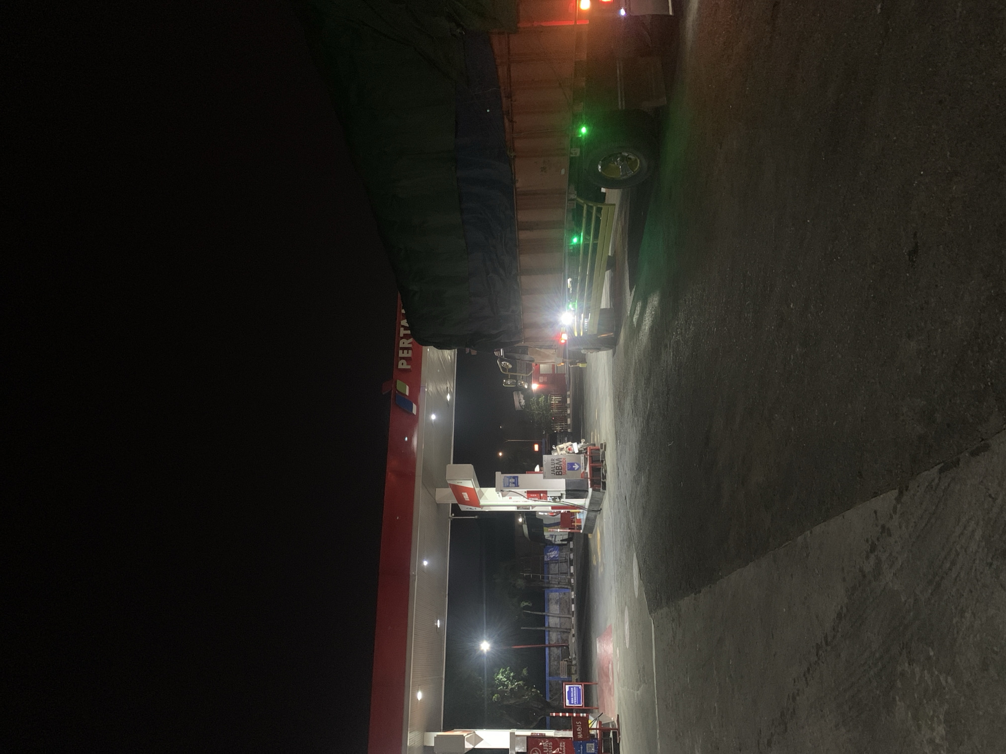 A nighttime view of a Pertamina gas station in Gambiran, Indonesia, with a large truck parked at a pump. The gas station canopy is lit with overhead lights, and the Pertamina logo is visible. The truck, covered with a dark tarp, has its side marker lights illuminated in green and red. The ground is asphalt with some tire marks. In the background, other vehicles and structures are dimly visible. The scene is illuminated by the artificial lights of the gas station and the vehicles.