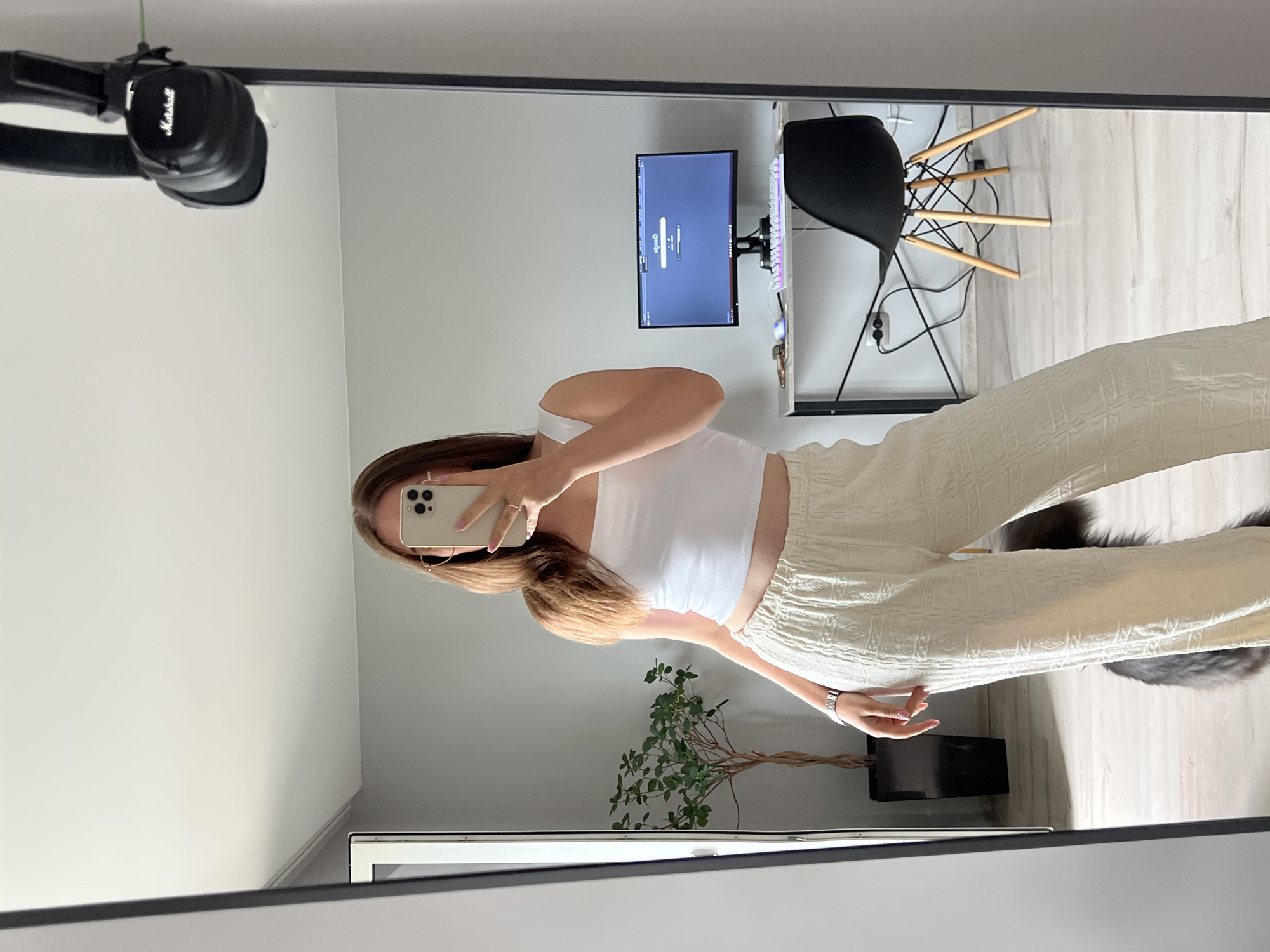 A full-length mirror reflects a young woman standing in a room, holding a smartphone to take a selfie. She is wearing a white tank top and cream-colored patterned wide-leg pants. Her hair is long and brown, and she is wearing glasses. In the background, a black desk with a computer monitor, keyboard, mouse, and keys is visible. A black Eames-style chair with wooden legs is positioned in front of the desk. A plant in a tall black pot stands to the left of the desk, and a pair of black Marshall headphones is hanging on the mirror's frame. The room appears to be a home office or living space, with light gray walls and a light-colored wooden floor. The scene is likely located in Ternopil, Ukraine, based on the provided context.