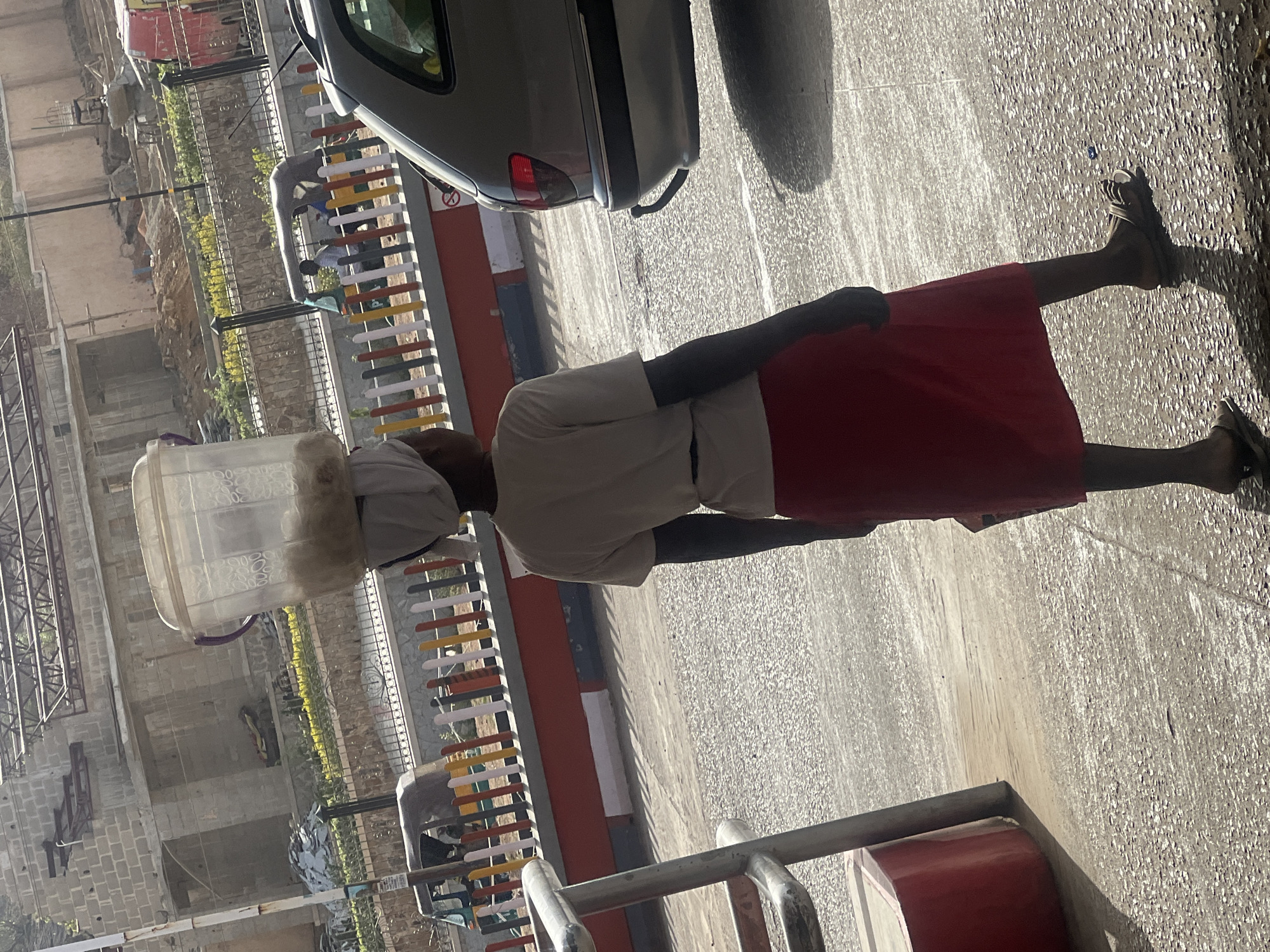 The image captures an individual, seen from behind, walking or standing on a paved surface under bright daylight in what appears to be an urban or semi-urban setting in Jos, Nigeria. The person, likely an adult, is skillfully balancing a large, translucent plastic container, possibly a water dispenser bottle, on their head, which is covered with a light-colored cloth. They are dressed in a plain, light-colored short-sleeved top and a long, flowing red skirt or wrapper, with sandals on their feet.
Strong sunlight casts distinct shadows on the ground, indicating a sunny day. In the midground, a silver sedan is partially visible, parked or moving near a colorful fence made of vertical slats in hues of white, yellow, red, grey, and black. Beyond the fence, the background reveals various structures, some appearing to be under construction with exposed concrete blocks and metal frames, alongside some green vegetation. The ground in the foreground is a textured, pebbled concrete surface. The scene depicts a moment of daily activity, highlighting a traditional method of carrying goods. No discernible text is visible in the image.