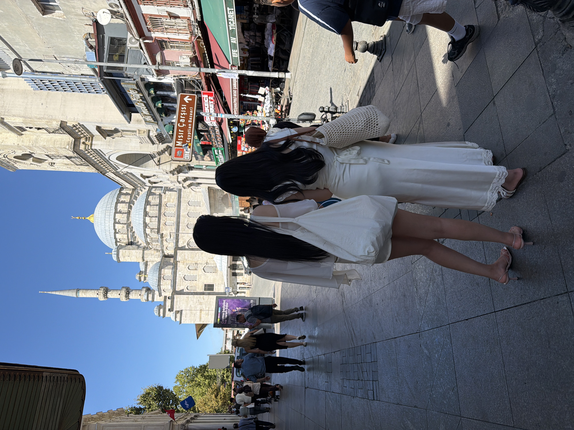 Two young women with long black hair are walking away from the camera on a paved street in Istanbul, Turkey. The woman on the left is wearing a short white dress and a sheer white top with a large white tote bag over her shoulder. She is wearing pink heeled sandals. The woman on the right is wearing a long white dress with lace trim at the hem and a cream-colored straw bag over her shoulder. She is wearing white sandals.
In the background, a large mosque with a prominent minaret and dome is visible under a clear blue sky. Shops with awnings line the street, and a street sign in Turkish reads "Mısır Çarşısı (Egyptian Bazaar)". Other people are walking on the street and sidewalk. The environment suggests a busy commercial area in Istanbul.