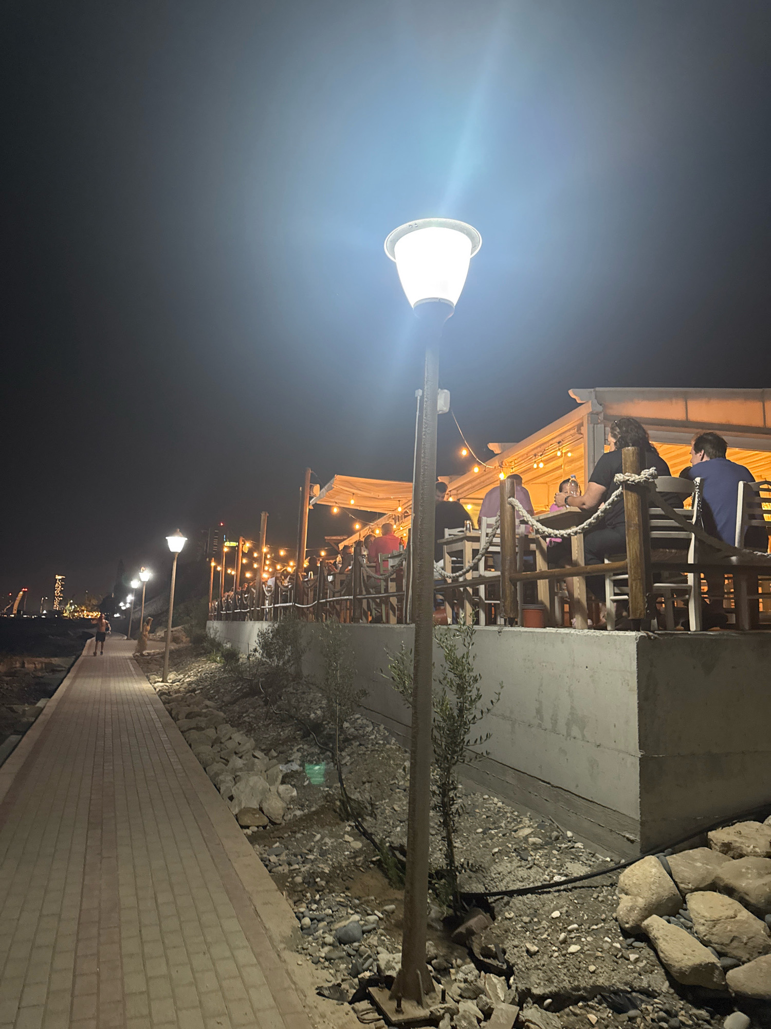 The image shows a nighttime scene at a beachfront restaurant in Agios Tychona Community, Cyprus. A paved walkway runs along the left side of the frame, illuminated by streetlights. In the background, a restaurant with a covered outdoor seating area is visible, adorned with string lights. Several patrons are seated at tables, some of whom appear to be dining and conversing. The area around the restaurant is rocky, with some sparse vegetation. In the distance, city lights and a tall building can be seen. The overall ambiance is one of a casual evening gathering.