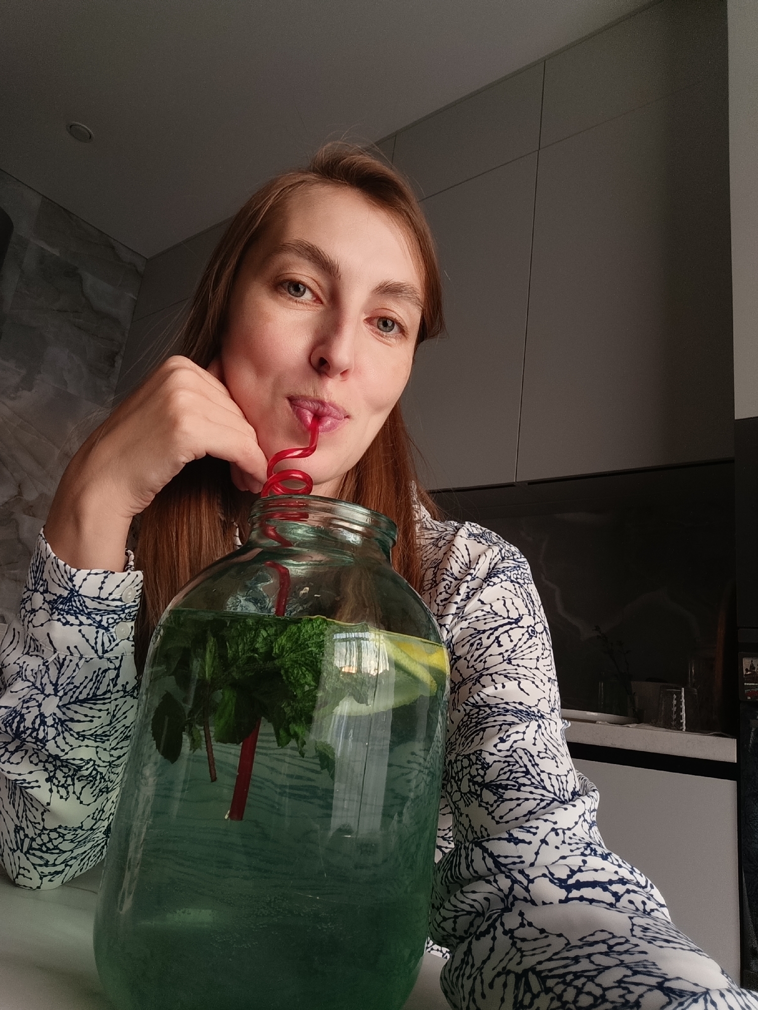 A medium shot captures a woman with reddish-brown hair drinking from a large glass jug filled with green liquid, mint, and lemon slices. She is positioned to the left of the jug, holding her chin with her left hand and a red, coiled straw in her mouth. The woman is wearing a white blouse with a blue abstract pattern. The background features a kitchen setting with gray cabinets and a marble-patterned wall. The lighting suggests it is daytime. The image is an indoor scene in a domestic environment, likely a kitchen. There is no visible indication of the specific location in Chaikovsky, Russia.