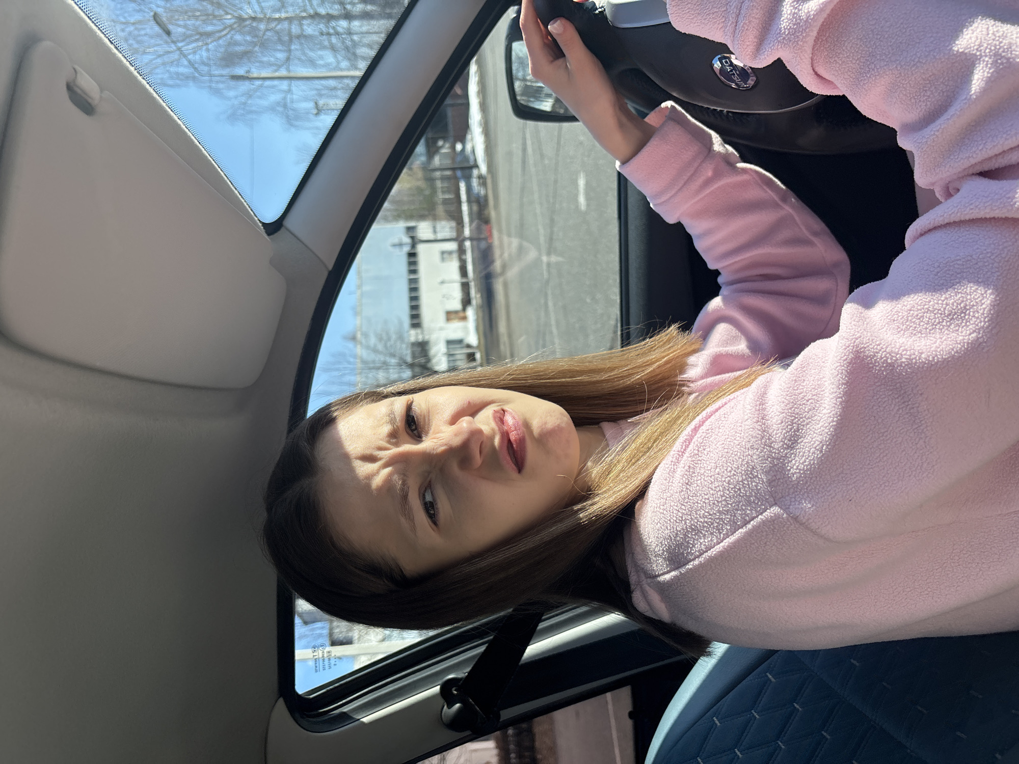 A young woman is sitting in the driver's seat of a car, with her hands on the steering wheel. She is wearing a pink sweater. The interior of the car includes blue quilted seats and a gray sun visor. Through the car windows, buildings are visible, suggesting an urban environment. The steering wheel has a Datsun logo, indicating the car model. The location is Chaikovsky, Russia.