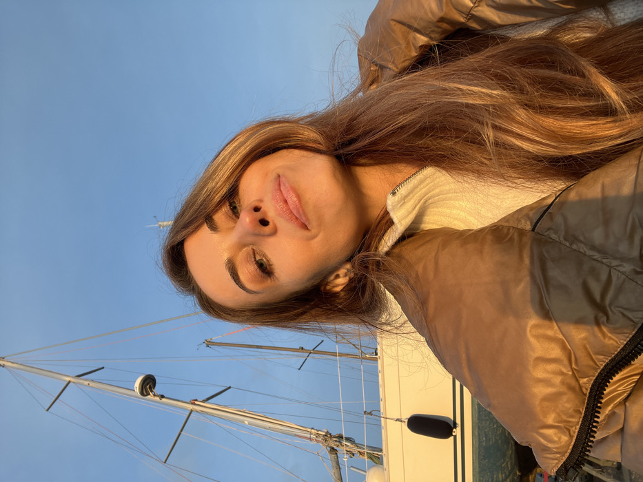 A woman with long blonde hair, wearing a tan jacket and a white sweater, poses for a selfie. Her face is angled, showing off her eyes and lips. Behind her, the blue sky contrasts with the mast and rigging of a sailboat, suggesting she's on a boat or near the water. The golden hour lighting casts a warm glow on her and the boat.