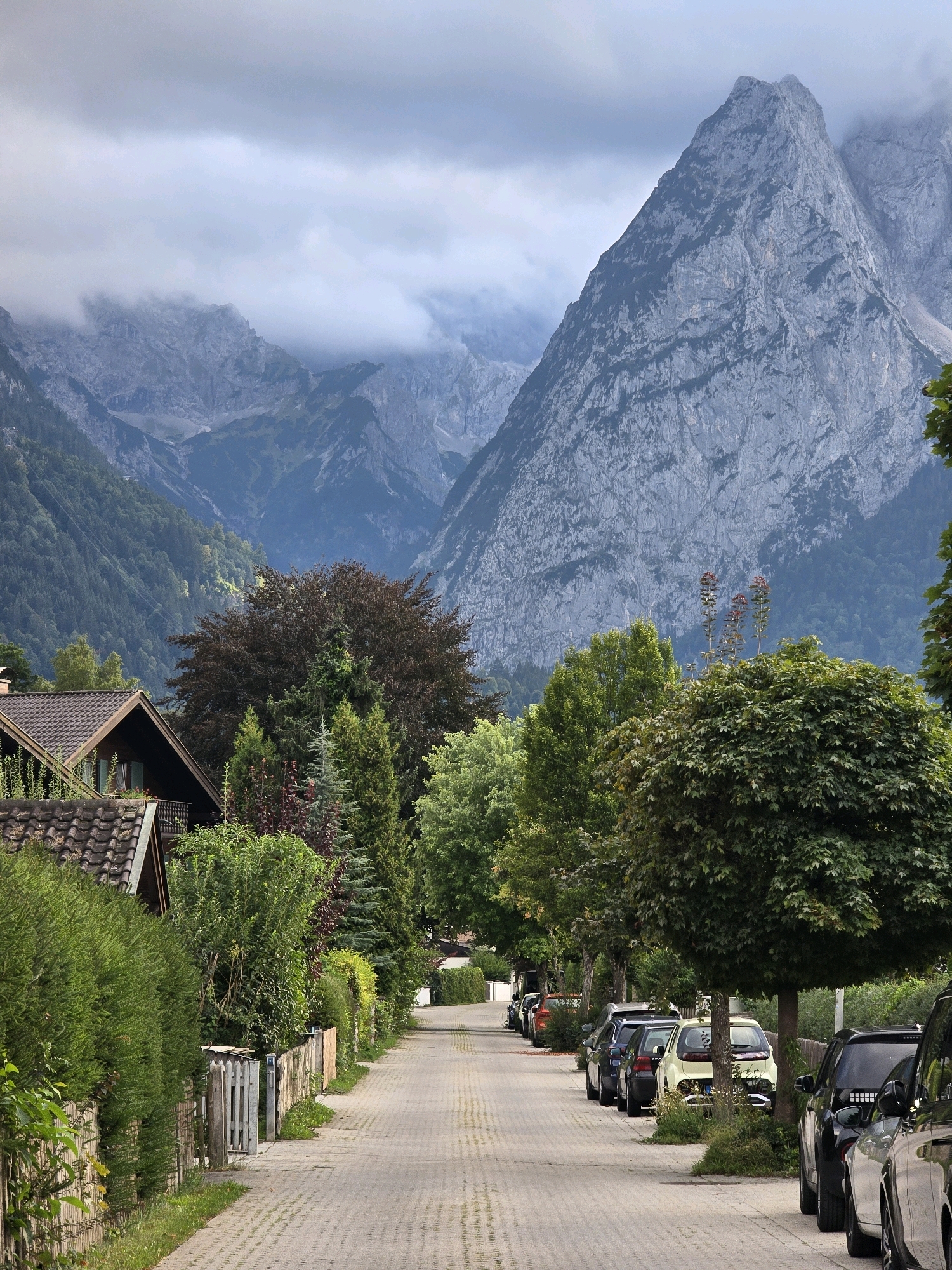 The image depicts a street in Garmisch-Partenkirchen, Germany, with a prominent mountain range in the background. The street is paved with brick-like stones and lined with houses on the left and trees on the right. Several cars are parked along the right side of the street. The houses are traditional Bavarian style, with dark tiled roofs and wooden facades. The mountain range is majestic and imposing, with jagged peaks partially obscured by clouds. Lush green trees and vegetation surround the houses and line the street, creating a picturesque and tranquil atmosphere.