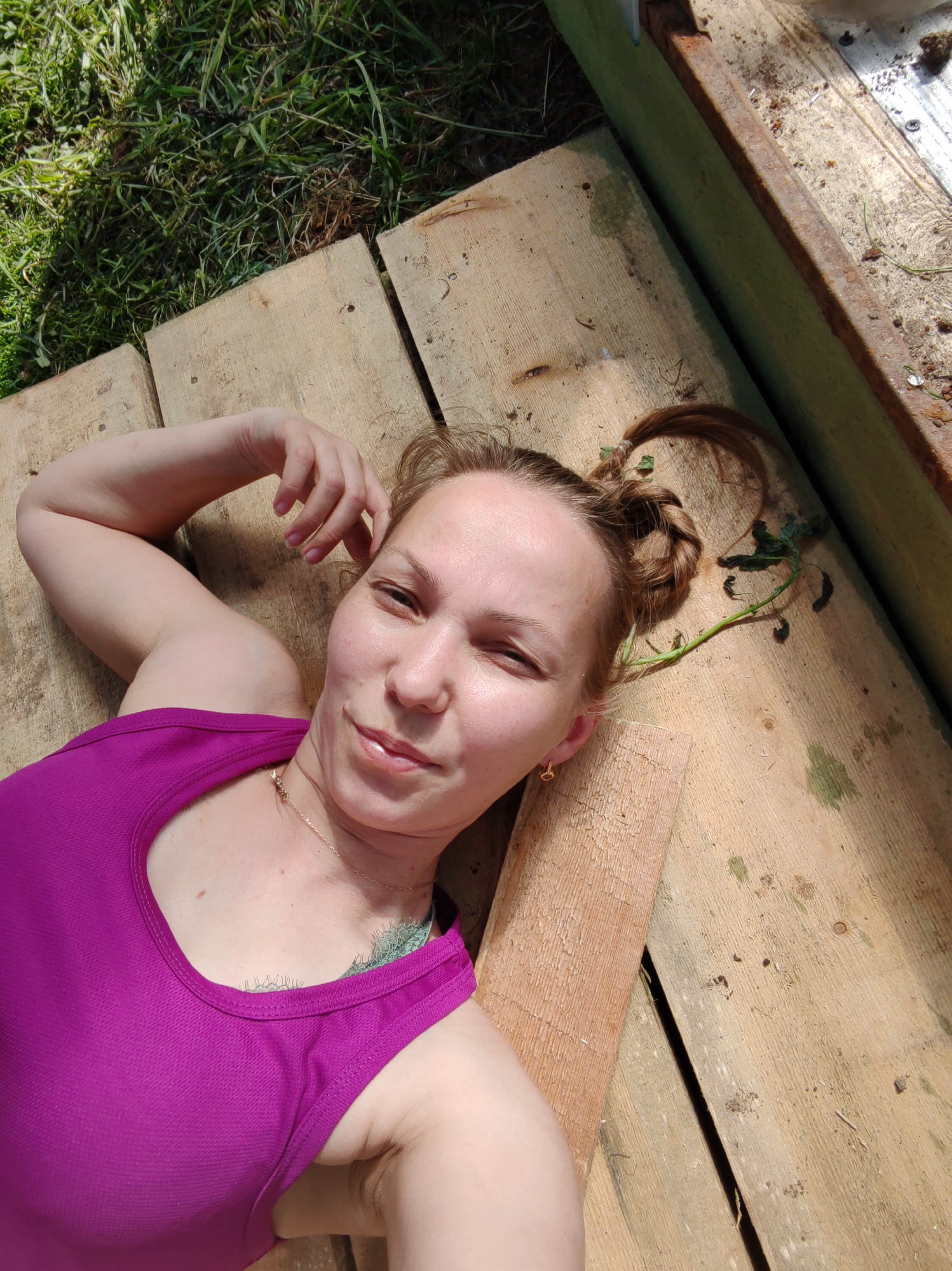 A woman is lying on wooden planks. She is wearing a magenta tank top and has her hair up in braided buns. Her left arm is bent, with her hand near her head. There is a plant with leaves on the planks next to her head. A green wooden structure is visible in the upper right corner. Grass is visible in the upper left corner. It is daytime, with sunlight illuminating the scene. The location is Чухлэм, Russia.