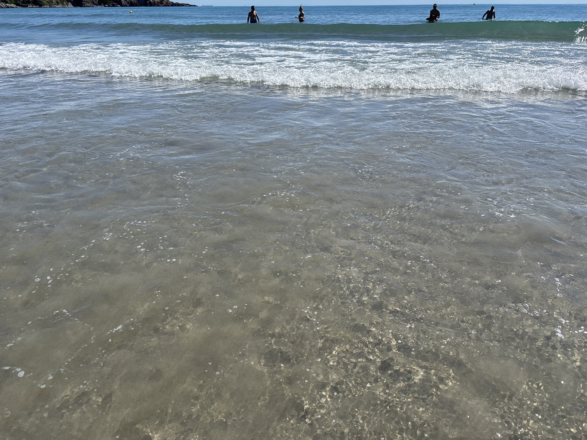 The image depicts a serene beach scene under a clear sky, suggesting a warm, sunny day. In the distance, a rocky coastline is visible, hinting at a coastal location. Several people are in the ocean, some standing in the shallow water and others further out. Their presence indicates recreational activity, likely swimming or wading. The water is calm with gentle waves lapping at the shore, creating a peaceful atmosphere. The sunlight reflects off the water's surface, highlighting the clarity of the shallow areas where the sandy bottom is visible. There are no discernible objects of interest or text in the image. The overall mood is tranquil and inviting, capturing a typical summer day at the beach.