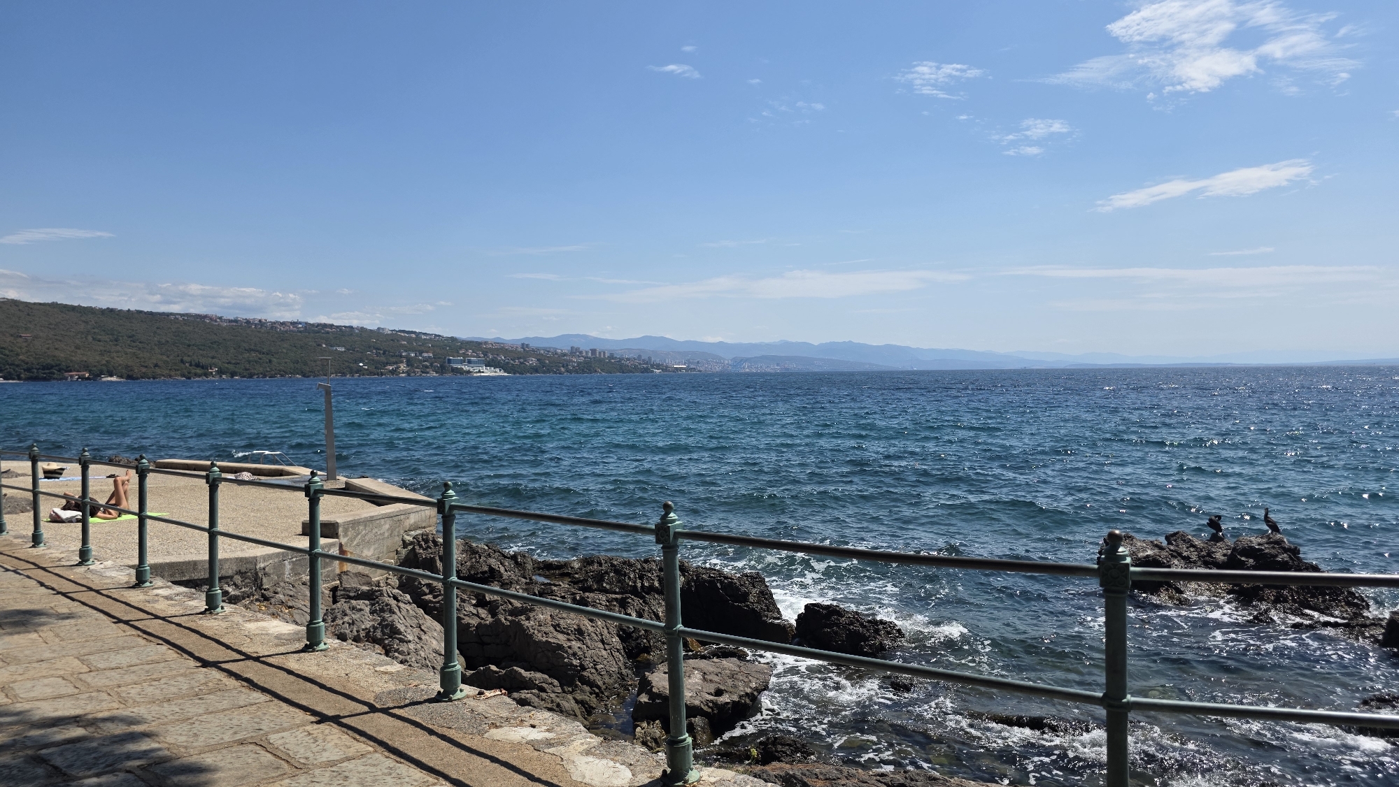 The image depicts a sunny day at the coast of Opatija, Croatia. In the foreground, a stone-paved walkway runs parallel to the sea, bordered by a dark metal railing. To the left of the walkway, a person is sunbathing on a green mat. The sea is a vibrant blue, with gentle waves lapping against the rocky shoreline. A distant coastline with green hills and a city skyline is visible across the water. The sky is clear blue with a few scattered white clouds. Several birds are perched on rocks in the water to the right of the image.