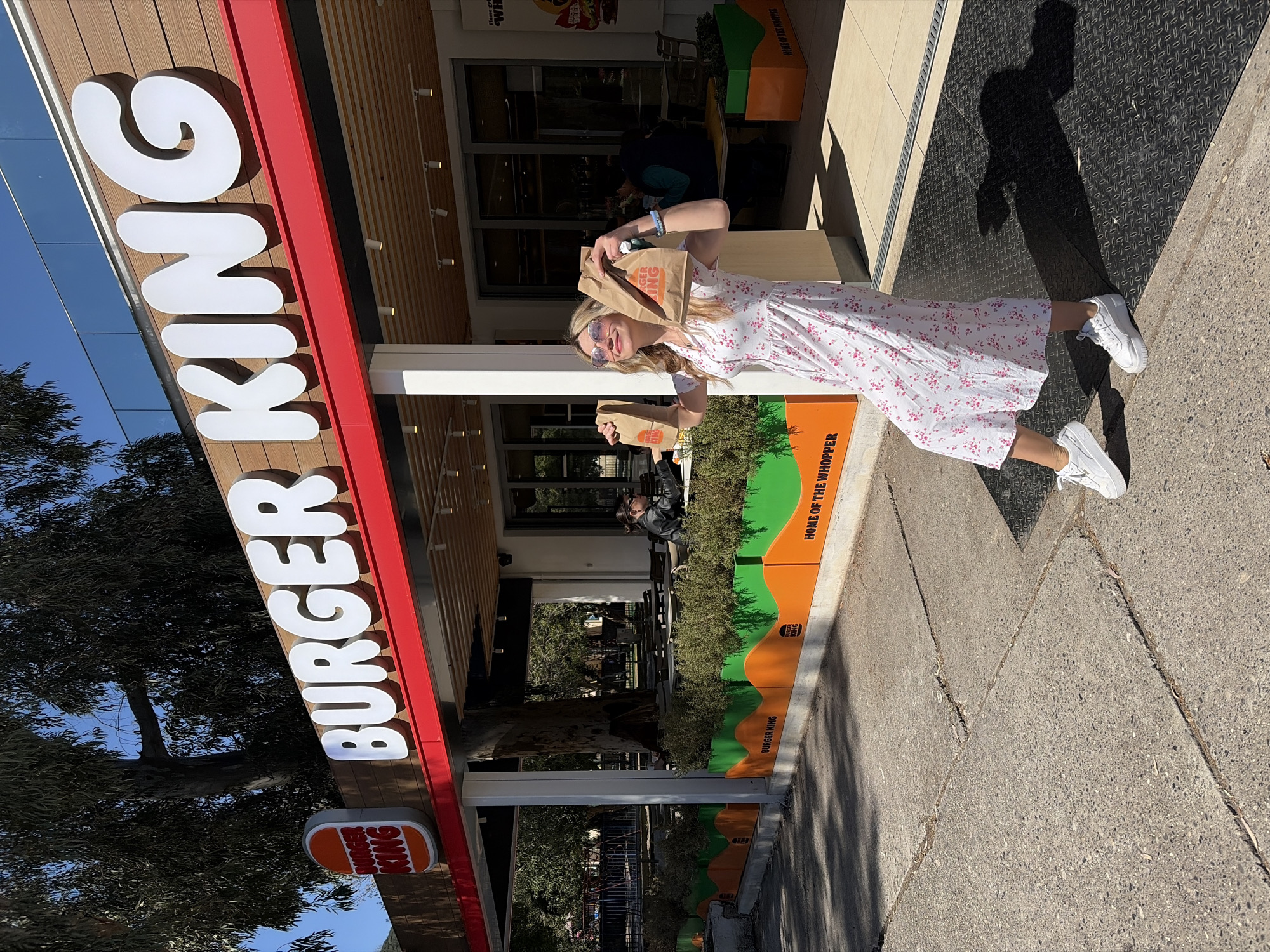 The image shows a woman in front of a Burger King restaurant. She is wearing a white dress with pink floral print and white sneakers. She is holding two Burger King bags up to her face and smiling. The Burger King logo is visible on the bags. The restaurant is located in Budva, Montenegro. A Burger King sign is visible above her head. The shadow of the photographer is on the ground.