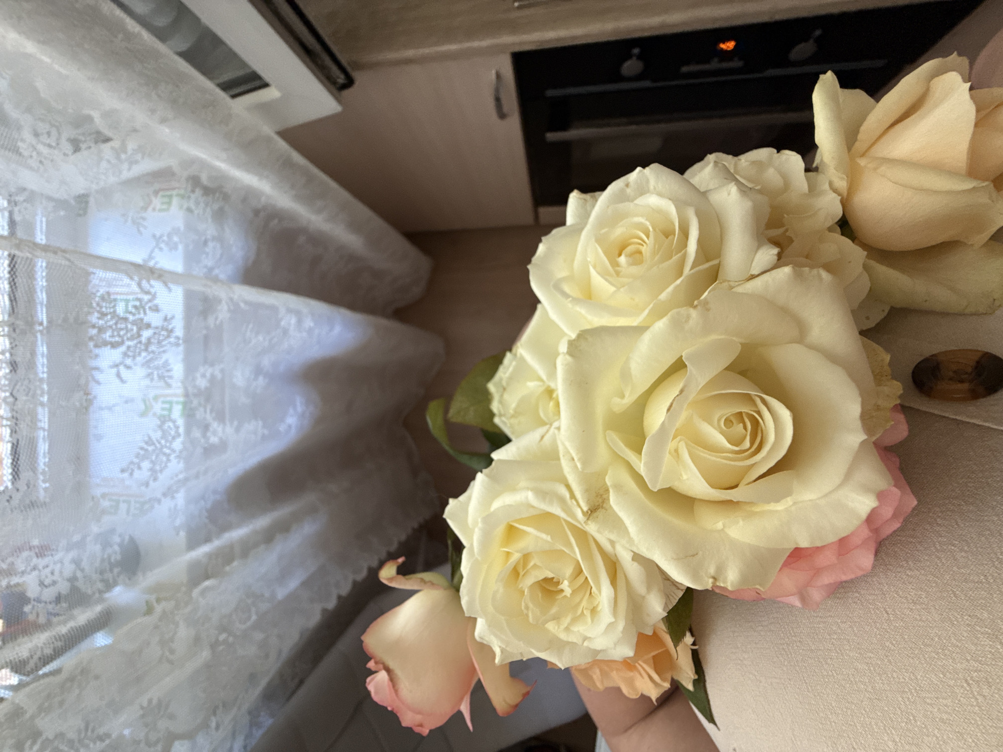 In an indoor setting, a bouquet of roses is held in someone's arms, positioned in the foreground. The roses are a mix of cream and pale pink hues, with their petals unfurling. Behind the flowers, a white lace curtain is draped across a window, filtering sunlight and casting subtle patterns. To the right of the curtain, a portion of a kitchen cabinet is visible, suggesting the room might be a kitchen or dining area. The background is softly blurred, with hints of the cabinet's wooden texture and a dark appliance, possibly an oven. The overall scene conveys a sense of domestic tranquility.