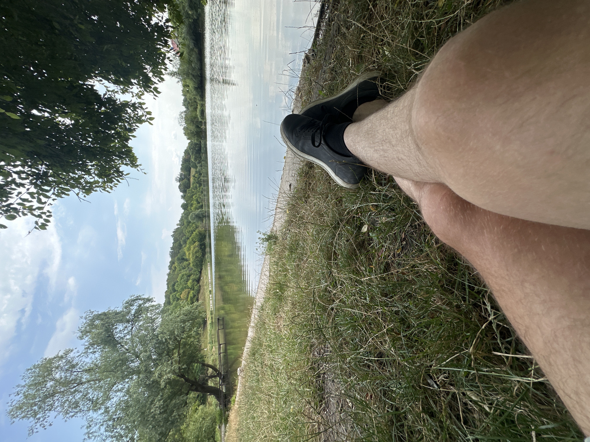 The image captures a low-angle perspective of a person's legs and feet resting on a grassy bank overlooking a body of water. The individual's bare legs, lightly tanned and covered in short hair, are crossed at the ankles, with black sneakers on their feet. The grass is dry and somewhat sparse, interspersed with patches of dirt and fallen leaves.
The background reveals a serene lake with calm waters reflecting the sky and the surrounding lush greenery. A tree with dense foliage stands prominently to the left, its branches casting shadows. Further in the distance, a line of trees and rolling hills forms the horizon under a partly cloudy sky. A wooden pier extends into the water on the left side of the frame.
This scene appears to be situated in a natural setting, possibly a park or a recreational area near Makiv, Ukraine, given the context of the provided information. The overall impression is one of relaxation and tranquility, with the subject enjoying a moment of repose by the water.