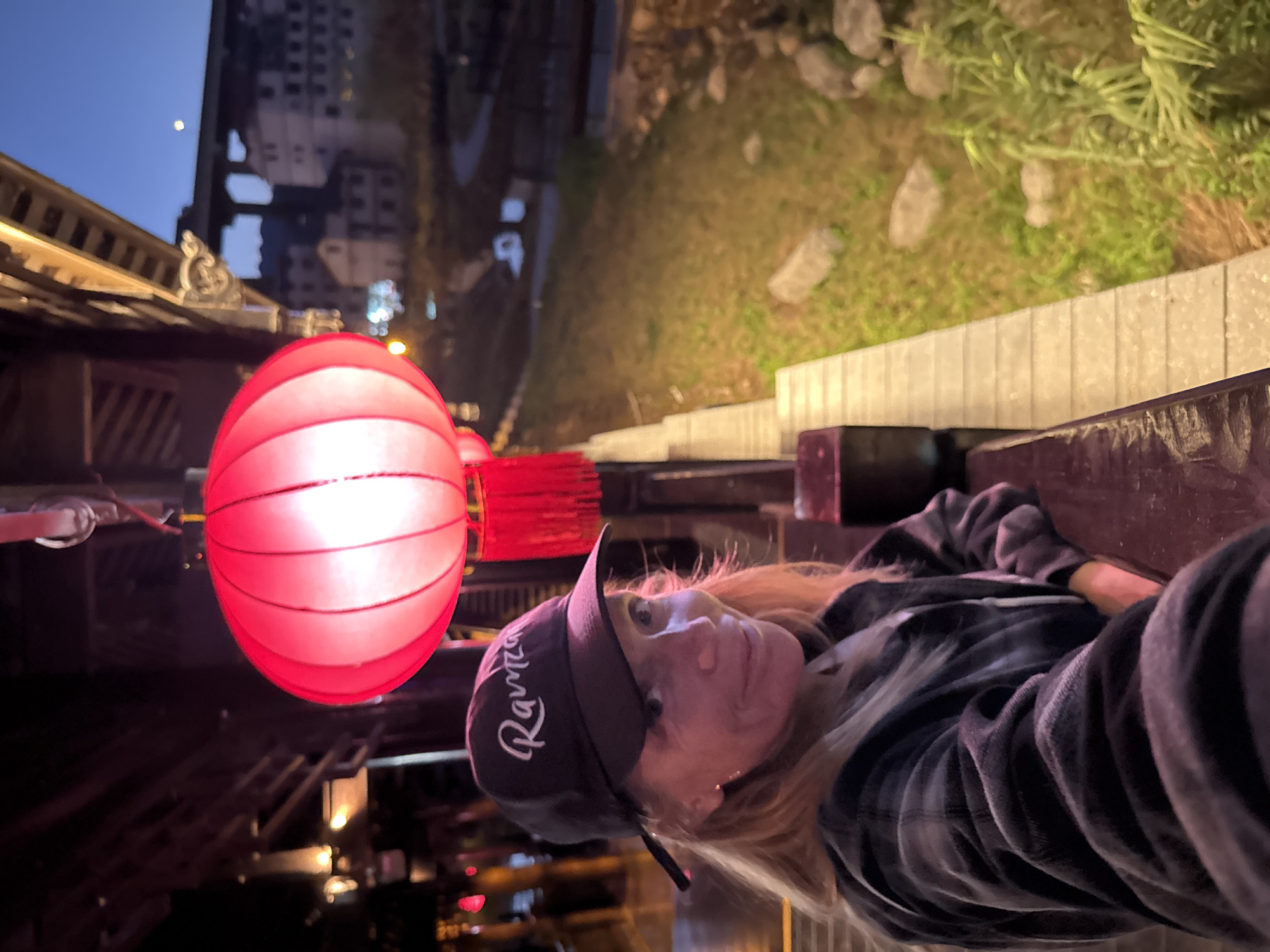 The photograph depicts a person wearing a dark-colored baseball cap with white cursive writing ("Rawmez") and a plaid shirt, leaning against a wooden rail or bench. A red Chinese lantern hangs nearby, illuminated and casting a soft glow. The background shows a street in Zhuoshui, China, with buildings, vegetation, and outdoor lighting. The sky is visible with a partial moon. The photograph appears to be taken at dusk or night.