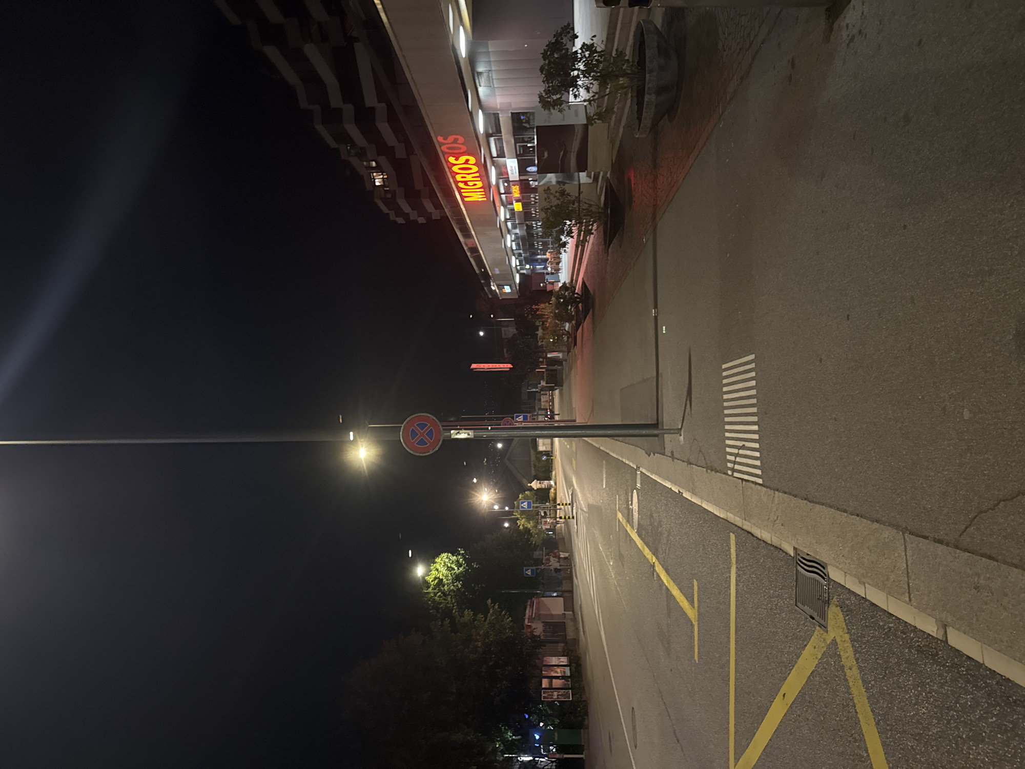 The image captures a street scene at night in Lugano, Switzerland. On the right side of the image, a Migros supermarket is illuminated by its prominent red neon sign. In front of the supermarket, a sidewalk is lined with planters containing shrubs. The street itself is paved, with yellow lane markings visible. A lamppost is centrally positioned in the foreground, casting light onto the scene. Traffic signs are present, including a red and blue circular sign with a diagonal red bar and an 'X' inside. The background features a multi-story building with several windows illuminated. Trees are visible on the left side of the street, and the sky is dark, indicating nighttime. The overall atmosphere is quiet and urban.