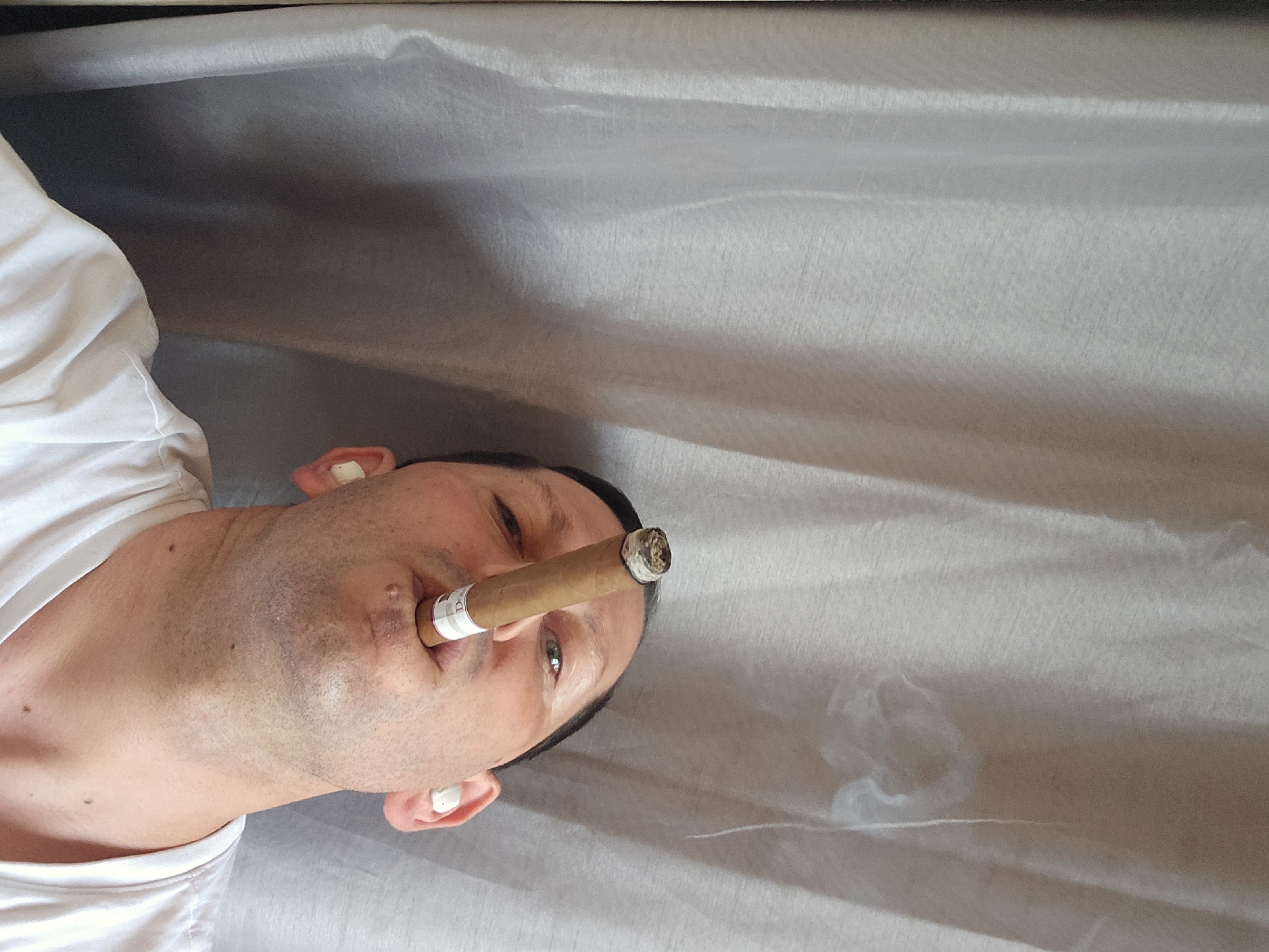 A man is smoking a cigar. The man is wearing a white shirt and is lying on a gray surface. Smoke is visible emanating from the cigar. The location is not identifiable as London, United Kingdom based on the visual information in the image.