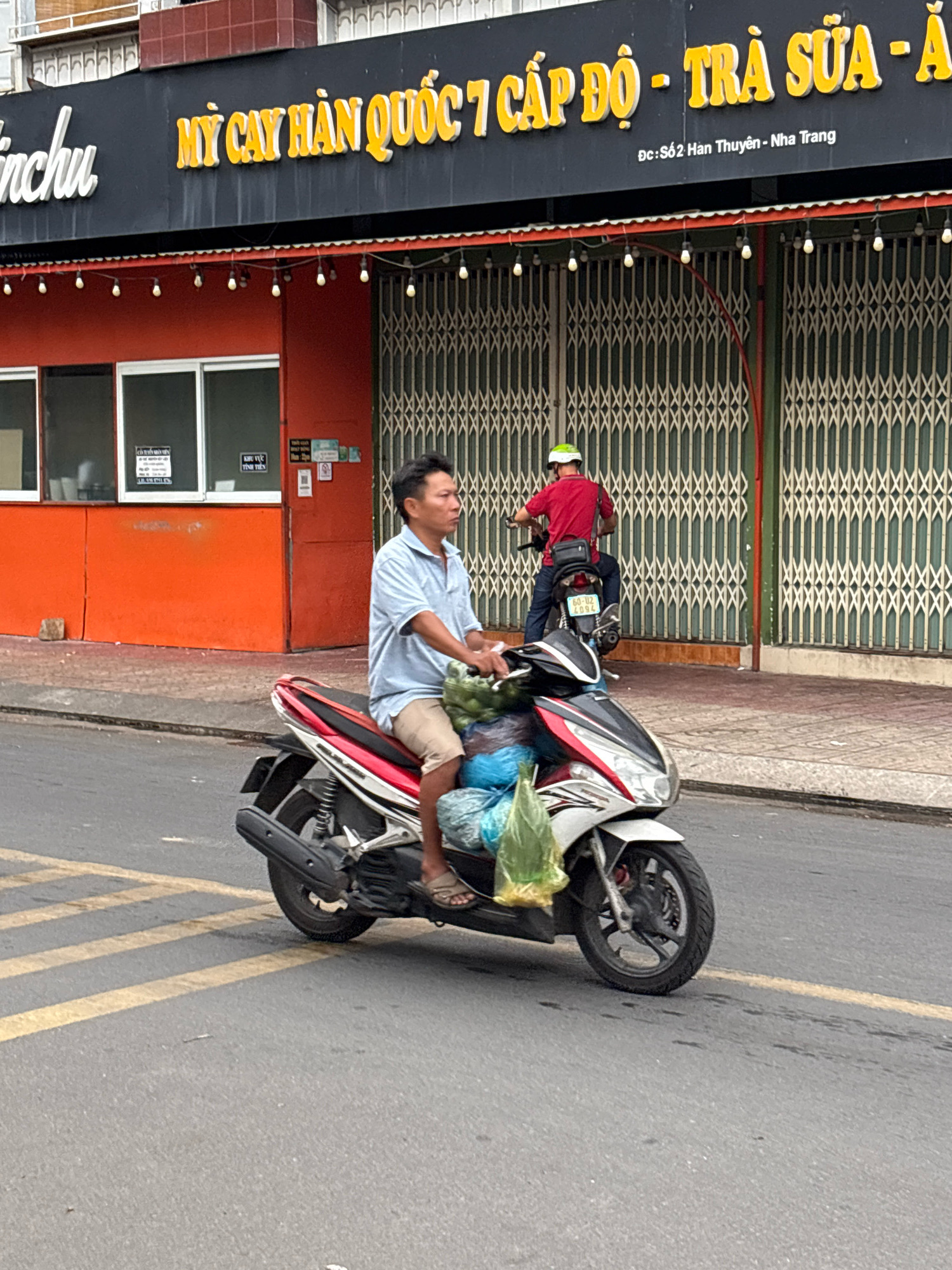 An outdoor, eye-level, medium shot captures a man riding a white, red, and black scooter on a paved city street in Nha Trang, Vietnam. The man, wearing a light blue collared shirt and beige shorts, has a concerned expression as he rides. He is carrying two large, clear plastic bags filled with green items, possibly vegetables or fruit, on the front of his scooter. The scooter has a license plate that reads "60-102 4094." In the background, a building with an orange facade and black awnings displays yellow Vietnamese lettering that translates to "My Cay Han Quoc 7 Cap Do - Tra Sua - A." Below this, in smaller lettering, is "Dc: So 2 Han Thuyen - Nha Trang." The building's entrance is covered by metal roll-up doors. Another person wearing a red shirt and a green helmet is visible on a scooter further down the street. The street has yellow lane markings.