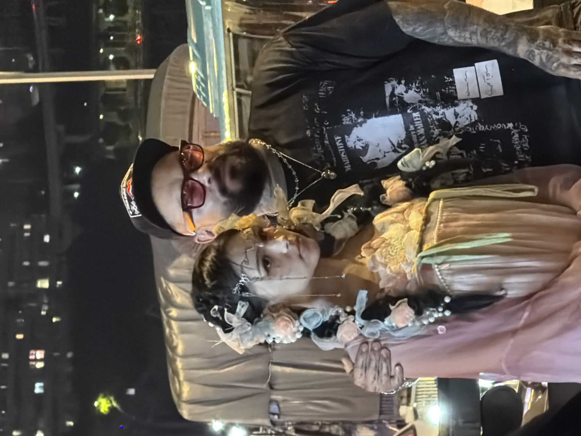 An adult male and an adult female are posed in front of a blurred urban nighttime background. The male, on the right, wears a black baseball cap, pink-tinted glasses, and a black t-shirt with a graphic print. He has a beard and tattoos on his neck and arms. His right arm is around the female's shoulders. The female, on the left, wears a pink dress and has an elaborate hairstyle adorned with flowers and pearls. Her head is tilted down and to the side, and her expression is somber. The environment appears to be outdoors at night, possibly in Yuzhong District, China, given the presence of city lights and structures in the background.