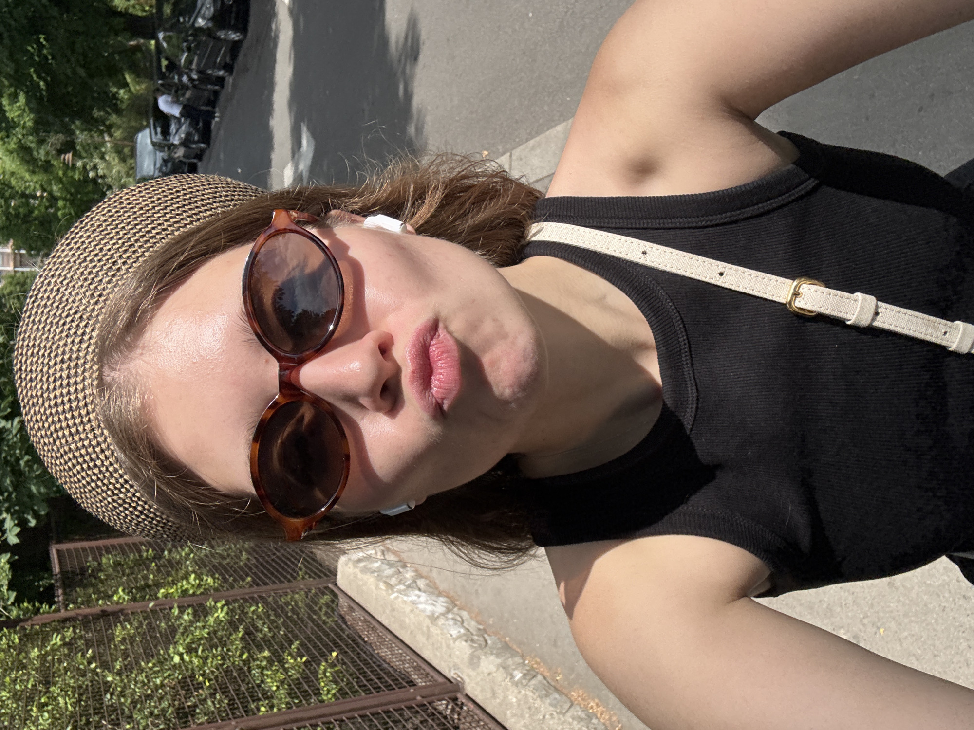 A woman is taking a selfie outdoors on a sunny day. She wears a black tank top, a brown patterned hat, and sunglasses. She has earbuds in her ears. A white strap crosses her body. The background includes a street, a fence, trees, and parked vehicles. The photo appears to be taken in an urban area. It is unclear if this photo was taken in Belgrade, Serbia.