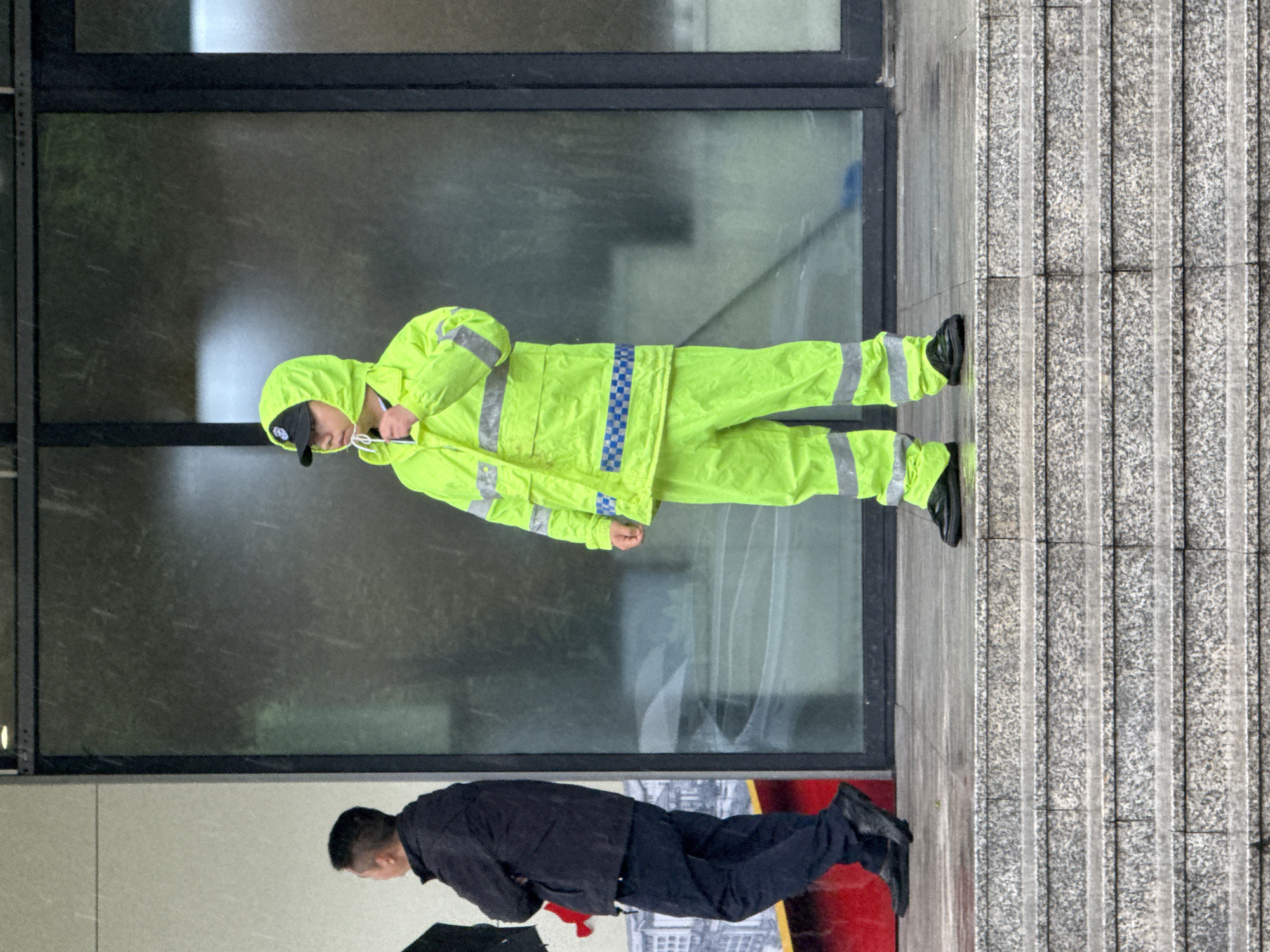 An outdoor, slightly desaturated, vertically oriented photograph captures two individuals in what appears to be an urban setting in Jiangjin, China.
The main subject, positioned centrally, is a person dressed in a full neon yellow-green reflective suit, consisting of a hooded jacket and pants. The suit features grey reflective strips on the arms and legs, and a distinctive blue and white checkered pattern across the chest. This individual wears a black baseball cap under the hood and is looking downwards, holding what appears to be a small white object or device with visible white strings in their hands. They are wearing black shoes.
Partially visible in the lower left portion of the image is another person, seen from the waist down, wearing a dark jacket and dark pants, also with black shoes.
The background features a large, dark-framed glass window extending across the upper and middle sections of the image, reflecting some indistinct shapes and light sources. To the right, a textured wall or floor surface, possibly made of concrete or granite, displays vertical grooves. The overall lighting suggests an overcast day or an indoor area with soft, diffused light. No visible text is present.