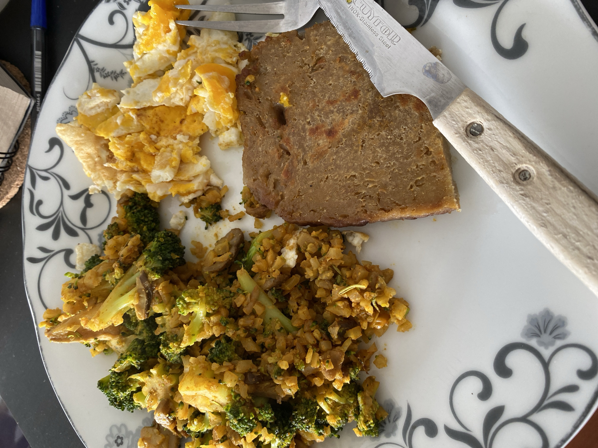 A plate of food sits on a patterned white plate. The food consists of scrambled eggs, a slice of meatloaf, and a mixture of broccoli and rice. A fork and knife rest on the plate, with the knife partially cutting into the meatloaf. The background is a dark surface with a pen, notebook and coaster. I have no information to suggest the photo was taken in the city of Gijón/Xixón, Spain.