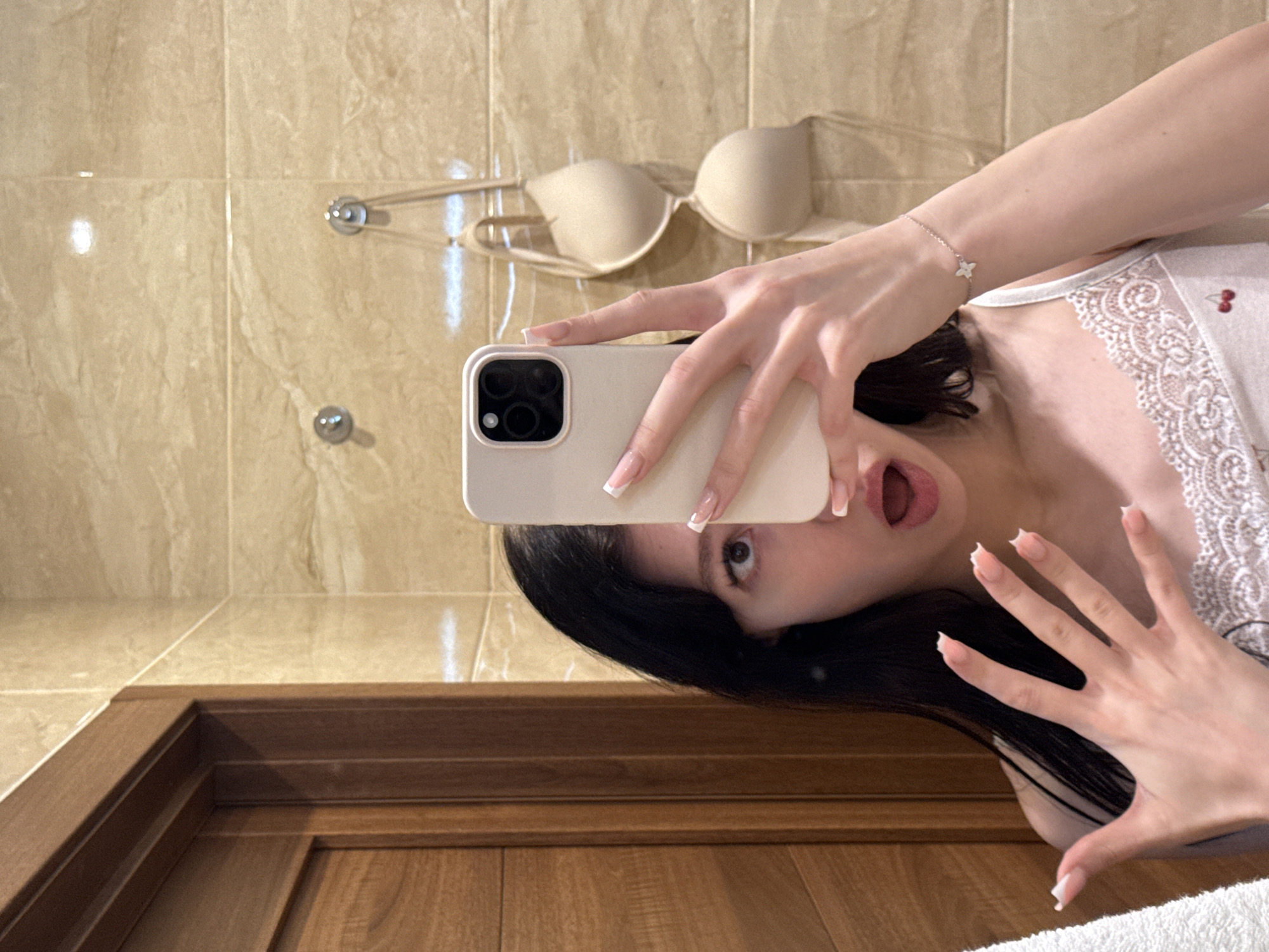 A young woman with long, dark hair is taking a mirror selfie in a bathroom. She is wearing a white lace-trimmed tank top with a cherry pattern. Her left hand is raised, holding a smartphone in front of her face, with her mouth open in a surprised expression. Her right hand rests on her arm, showing a bracelet with a small star charm. Behind her, a beige bra is hanging on hooks on a tiled wall. The bathroom also features a wooden doorframe on the left. The image is taken indoors, likely in a residential bathroom. There is no information in the image to suggest a location in Khimki, Russia.