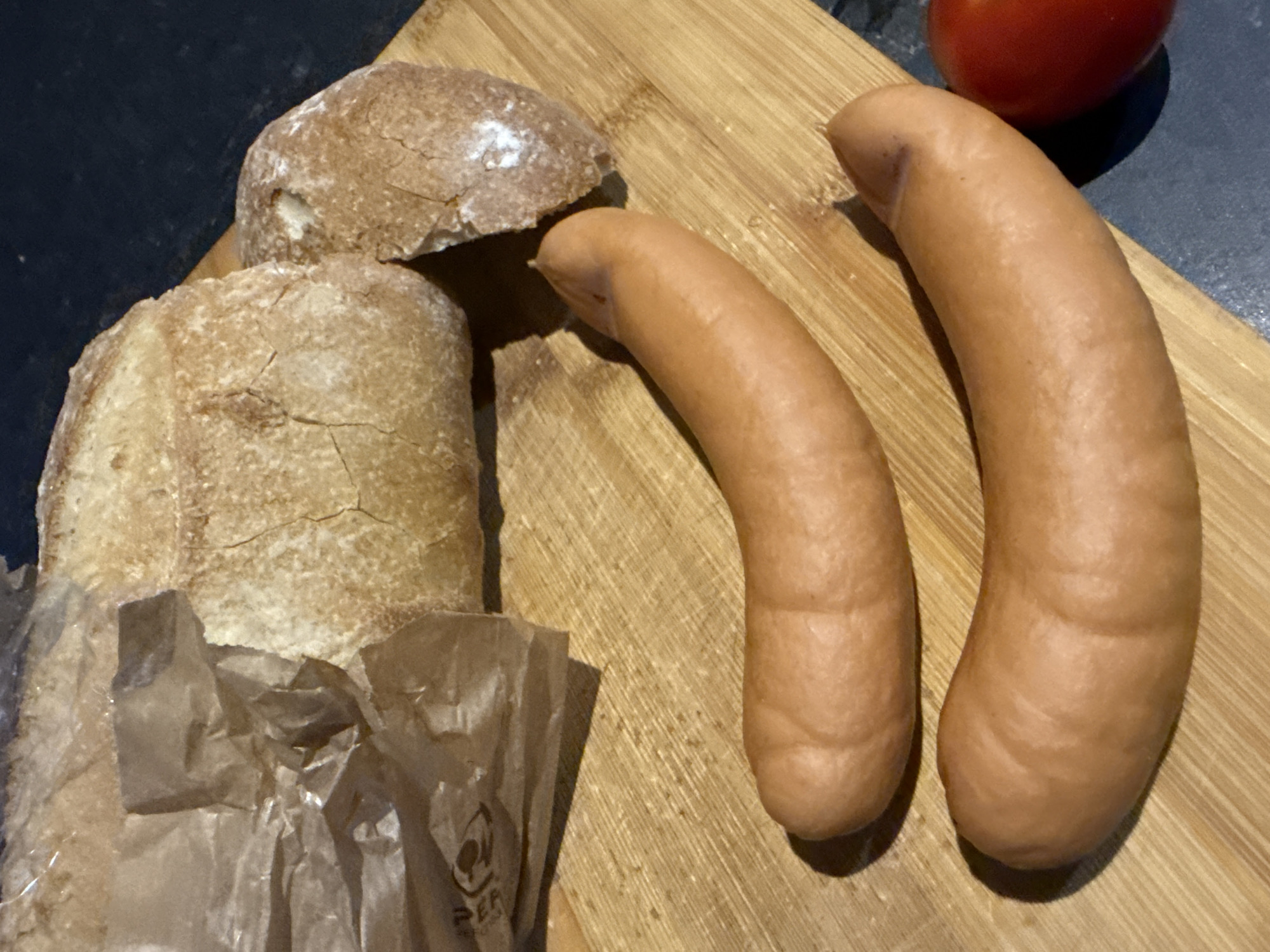 Two sausages lie on a wooden cutting board next to a partially opened paper bag and two pieces of bread. A single red tomato is visible in the bottom right corner. The image is taken from a top-down perspective, with the background appearing dark. The lighting suggests an indoor setting. No people are present in the image, and there is no information that allows for the determination of its location in Great Yarmouth, United Kingdom.