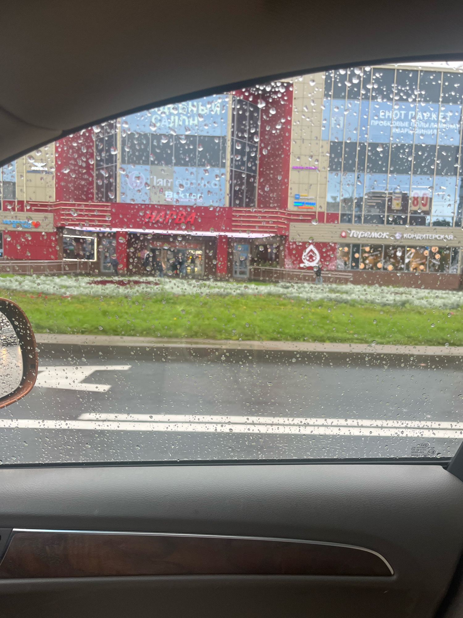 The image shows a view from inside a car, looking out through a rain-streaked window at a modern shopping center in Saint Petersburg, Russia. The building has a red and beige facade with large glass windows. Some of the signage visible on the building includes "салон" (salon), "ЕНOT ПАРКЕТ" (Enot Parket - flooring store), and "Теремок КОНДИТЕРСКАЯ" (Teremok Confectionery). There are people walking inside and outside the building, and a green lawn with white and red flowers in front of it. The road in front of the shopping center is wet, indicating recent rain. A car's side mirror is visible on the left side of the frame. The interior of the car is also visible at the bottom of the frame, showing a gray door panel with a wood-grain trim.