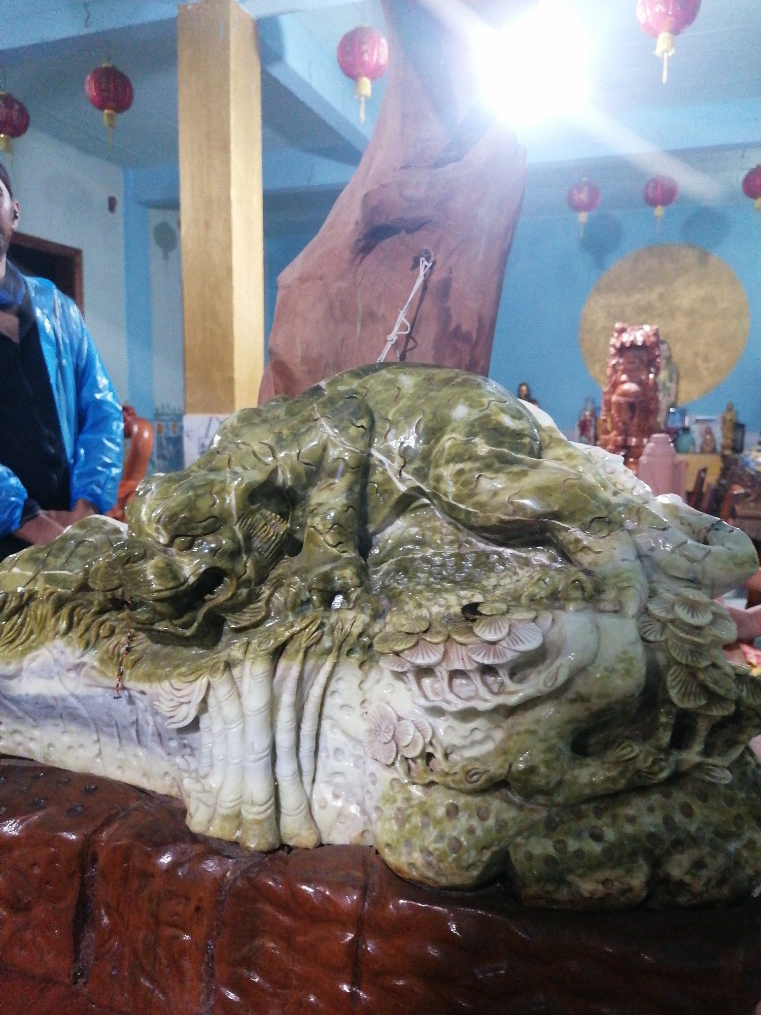 The image features a large, intricately carved sculpture made of a mottled green and white stone, likely jade or a similar material, positioned centrally on a dark, reddish-brown textured base. The carving depicts a detailed mythical creature, possibly a dragon or a similar legendary beast, with scales, a prominent head, and stylized elements resembling feathers or fins.
On the left side, a person is partially visible, wearing a shiny blue jacket over a black garment. Only their shoulder, arm, and part of their head are in frame; their face is not clearly seen.
The background reveals an indoor setting with light blue walls. Several red, spherical Chinese-style lanterns hang from the ceiling. A tall, golden-colored pillar stands vertically in the mid-ground. Further back on the right, a large golden circular decoration is affixed to the blue wall, and numerous smaller, reddish-brown statues and carvings are arranged on shelves or tables. A very bright light source, possibly an overhead light or a window, creates a strong highlight in the upper center of the image.
The scene suggests a gallery, shop, or cultural space where artisan carvings are displayed or sold. The presence of the lanterns and the style of the carvings are indicative of East or Southeast Asian cultural aesthetics, aligning with the given location context of Da Lat, Vietnam. There is no specific activity beyond observation or display evident, and no visible text. The bright illumination suggests it is likely daytime or a well-lit interior.
