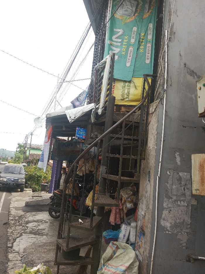 A narrow, dark metal spiral staircase hugs the side of a weathered building. Banners hang above the stairs, advertising beverages. Debris and goods clutter the base of the staircase, including sacks, plastic bags, and cloths. The building's exterior is worn, with peeling paint and visible cracks. A motorcycle is parked nearby, and a vehicle is on the adjacent road. Overhead, a tangle of wires crisscrosses the sky.