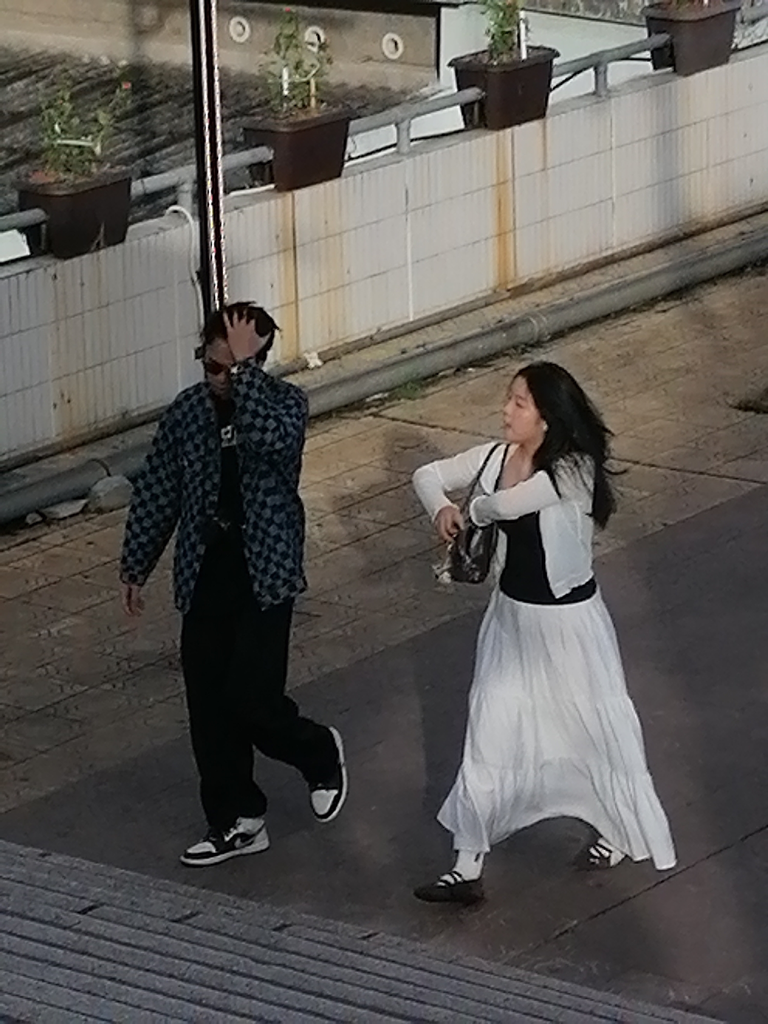 Two individuals are walking on a paved walkway in Da Lat, Vietnam, with potted plants and a tiled wall in the background. The individual on the left, a male, is wearing a black t-shirt, a checkered blue and black jacket, black pants, and black and white sneakers. He is touching his hair with his right hand and wearing sunglasses. The individual on the right, a female, is wearing a black top, a white long-sleeved cardigan, a long white skirt, and dark shoes with white socks. She is holding a dark handbag in her right hand and appears to be looking to her left. The walkway is made of grey stone slabs, and there are metal railings and pipes visible in the scene.
