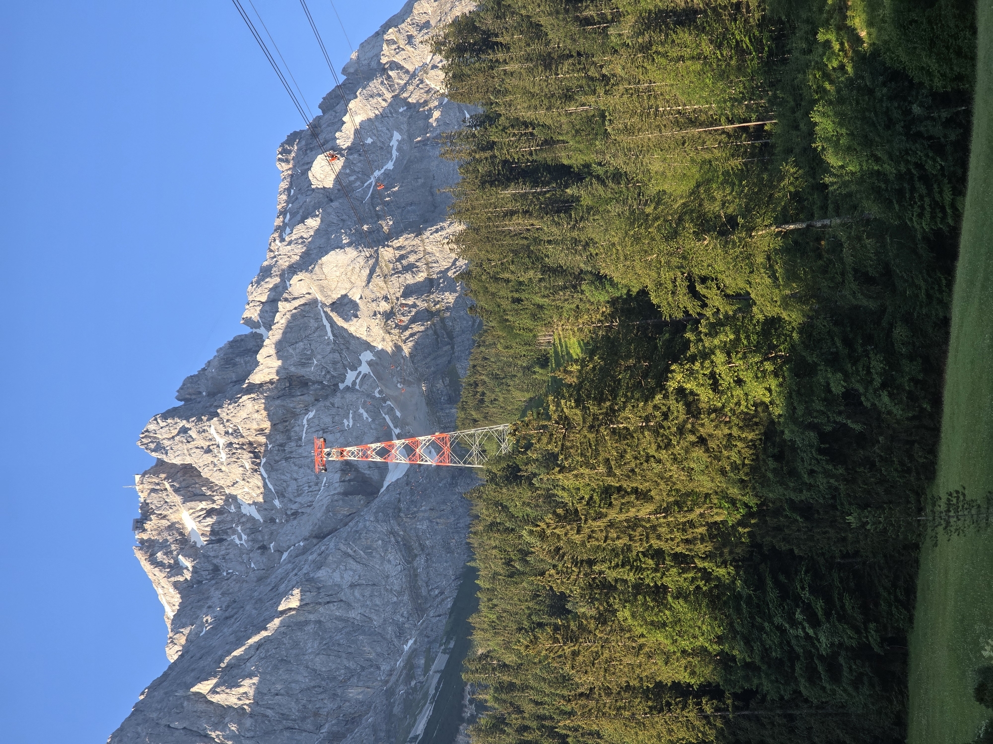 The image depicts a mountainous landscape with a clear blue sky. In the foreground, a dense forest of evergreen trees stretches across the frame. A red and white lattice tower stands prominently against the rocky mountain face, which is partially covered in snow. Several cable cars are visible ascending the mountain, indicating that a cable car system is in operation. The lighting suggests it is either early morning or late afternoon, with long shadows cast across the mountain. The overall scene conveys a sense of natural beauty and outdoor recreation. No visible text is present in the image. The location context provided is Grainau, Germany.