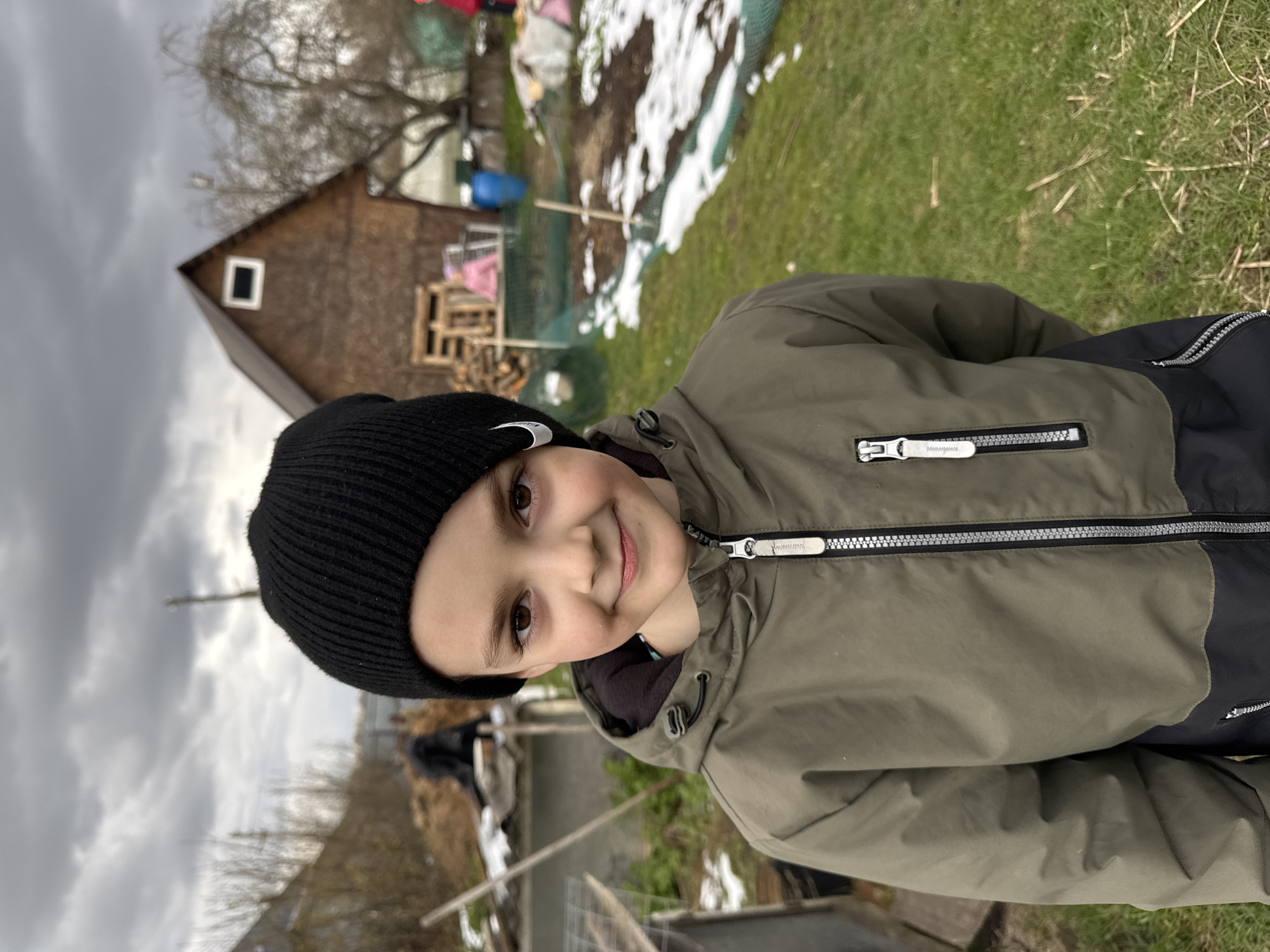A portrait-oriented outdoor photograph captures a young boy with light skin, brown eyes, and a gentle smile, looking directly at the camera. He wears a black ribbed knit beanie with a small white emblem on the left side, and a practical olive green jacket featuring black panels on the sides and sleeves. The jacket has prominent silver zippers with white pulls; one zipper pull on the left chest clearly displays the small white text "WWW.WWW" on a black background.
The setting appears to be a rural or semi-rural area, likely a backyard or homestead, in Раздолье, Russia. The background features an overcast, grey sky dominating the upper left quadrant. Below the sky, to the boy's upper left, a brown wooden house with a dark roof and a small white-framed window is partially visible. Bare tree branches are scattered throughout this area, suggesting a cold season.
To the boy's right and extending into the background, patches of snow lie on green grass and exposed earth, indicating a late winter or early spring day. Various outdoor items are discernible, including wooden pallets and a blue barrel. The boy is posing for the photograph in an outdoor environment, characterized by chilly weather and natural elements.