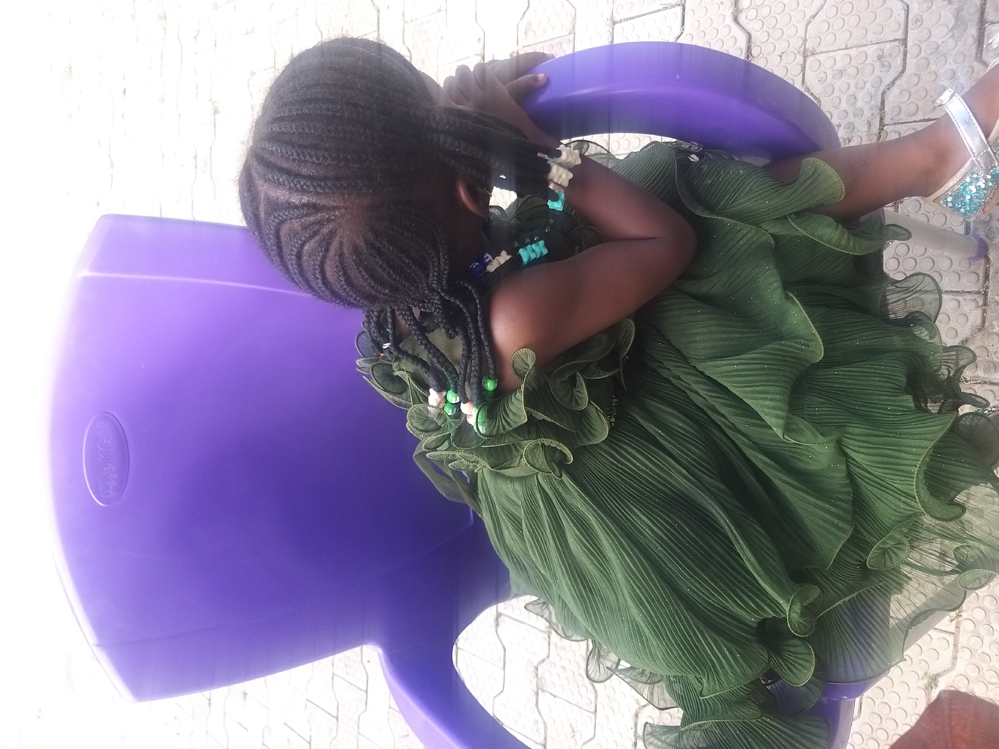 A young girl with dark skin and braided hair, adorned with green and blue beads, sits on a purple plastic chair. She wears a voluminous, dark green ruffled dress with glitter. Her head is bowed, resting on her arm, which is supported by the armrest of the chair. The chair is positioned outdoors on a tiled surface. The environment appears to be an urban setting, possibly a residential compound or a public space within Kano, Nigeria. The lighting suggests it is daytime.