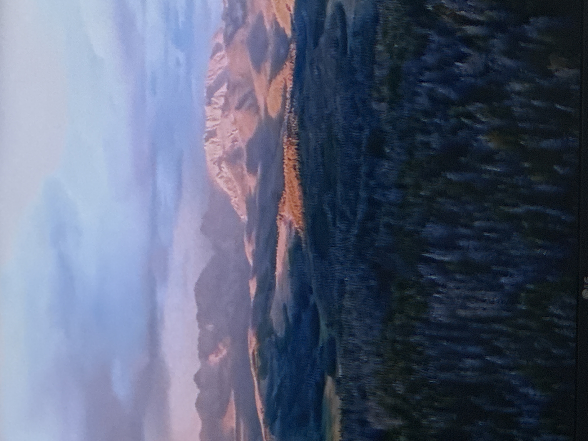 The image depicts a broad, mountainous landscape at dusk. The sky is a gradient of soft blues and purples, with scattered clouds. In the mid-ground, rolling hills are covered in dark green pine trees, with some patches of lighter, possibly autumnal vegetation. The sun, low on the horizon, casts a warm, golden-pink glow on the highest mountain peaks in the background, highlighting their rugged terrain. Shadows are long and deep, obscuring detail in the valleys. The foreground is dominated by a dense forest of dark trees, with a slightly lighter, more textured appearance suggesting the upper canopy. The overall impression is one of serene, natural beauty. There is no indication of the city of Sinyavino, Russia, in this image.