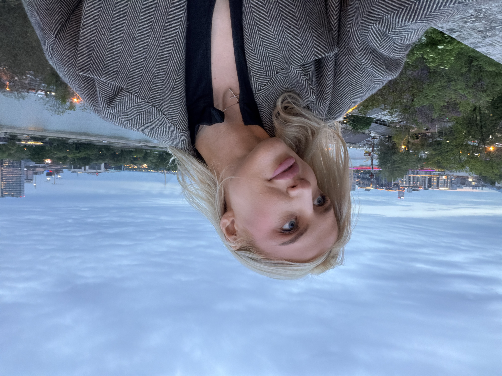 A portrait-oriented image shows a young woman at dusk or early night, positioned against an urban waterside backdrop. The subject is a young woman with blonde hair, blue eyes, and fair skin, looking directly at the camera with a slight pout. She is wearing a dark top underneath a grey and white herringbone pattern jacket, and a delicate silver-colored cross necklace is visible around her neck.
Behind her, a tranquil urban scene unfolds. A body of water, likely a river, stretches across the midground, reflecting the twinkling lights of the city. The riverbanks are lined with lush greenery and some urban infrastructure. In the background, a cityscape rises, featuring numerous buildings illuminated against the evening sky. The sky above is characterized by soft blues and greys with layered clouds, indicative of either dusk or dawn. The overall impression is one of a serene urban setting during the transition from day to night. The photograph appears to be a close-up selfie or portrait. The context provided indicates the location is Belgrade, Serbia, consistent with the river and city skyline visible. No discernible text is present in the image.