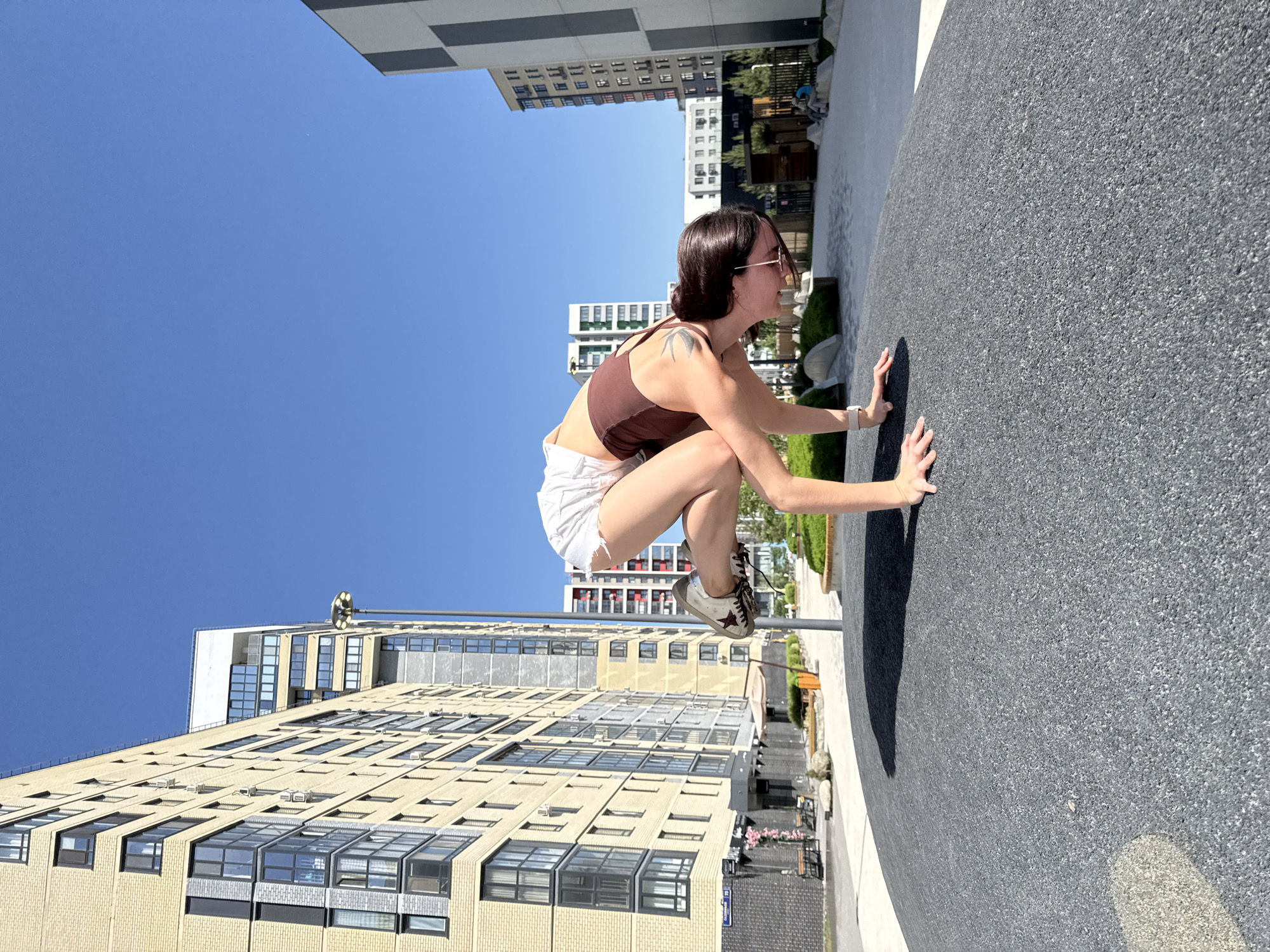 A young woman with dark hair and sunglasses is captured mid-air, seemingly hovering parallel to the ground, in an urban setting in Tyumen, Russia. She is wearing a brown tank top and white shorts with tattered edges, sneakers, and a watch. Her hands are outstretched, touching the dark grey, granular surface, suggesting she might be performing a handstand or a levitation trick for a photo.
The scene is set against a backdrop of modern high-rise buildings with yellow brick facades and numerous windows. The sky is clear and blue, enhancing the daylight. Other architectural details, such as a lamp post and greenery, are visible, helping define the urban landscape.