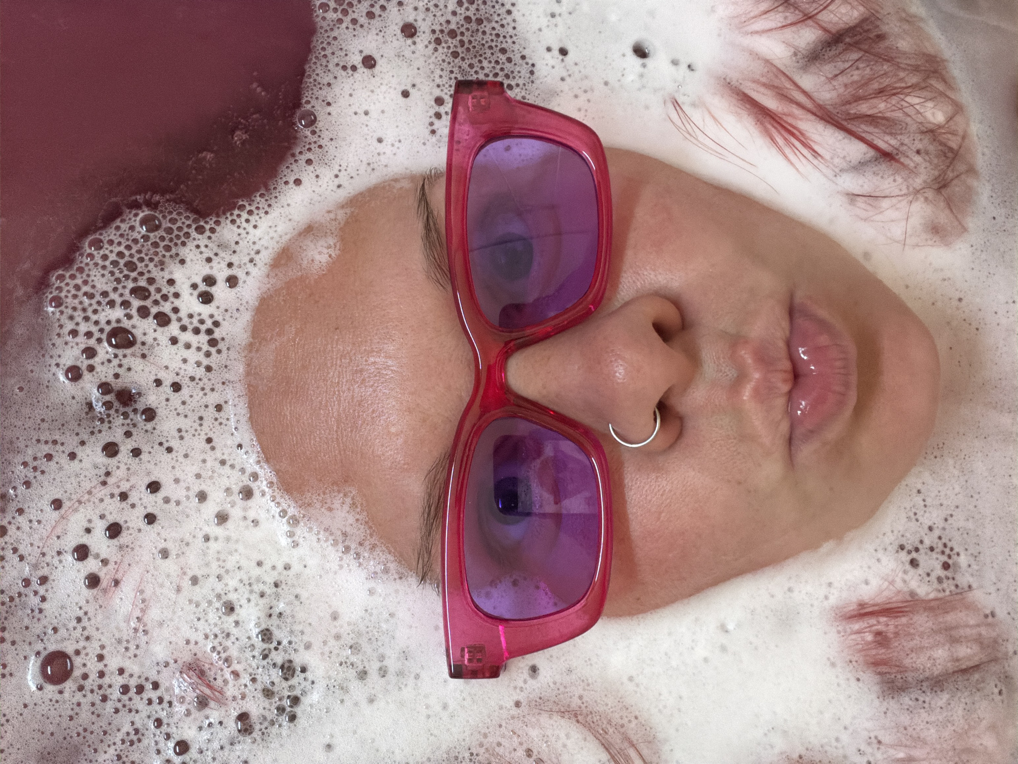 The image is a close-up, overhead shot of a person's face submerged in a pink, bubbly bath. The person is wearing bright pink sunglasses and a silver nose ring. Their lips are pursed, and their eyes are visible through the purple lenses of the sunglasses. Red hairs are scattered in the bubbles around their face, and a darker pink liquid is visible in the upper right corner, suggesting it is a bath bomb or colored bath water. There is no discernible city or location in the image.