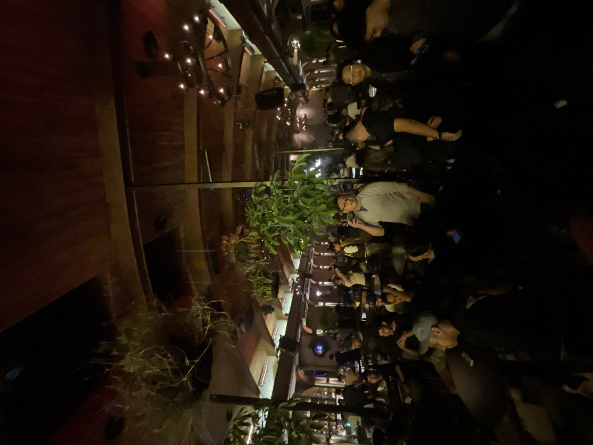 The image depicts an indoor gathering at a dimly lit establishment in North Jakarta, Indonesia. The scene is characterized by a low-hanging, slatted wooden ceiling adorned with hanging plants and decorative light fixtures. In the foreground and midground, numerous individuals are visible, clustered around tables and standing. The lighting is subdued, casting long shadows and creating a somewhat intimate atmosphere. Several people are engaged in conversation, while others appear to be observing their surroundings. The overall environment suggests a social setting, possibly a restaurant, bar, or event space, with an emphasis on relaxed social interaction.
