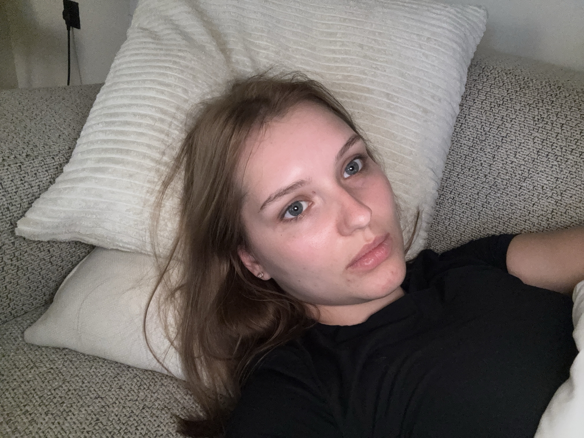 A young woman is lying on a couch, propped up by pillows. She has light brown hair and blue eyes, and she is looking directly at the camera. She is wearing a black t-shirt. The couch is upholstered in a textured gray fabric, and there are white pillows behind her. The lighting suggests it is evening or indoors. The woman's expression is neutral, with slightly parted lips. There are no other people or objects prominently visible in the image. The scene appears to be a casual, indoor setting, likely a living room. There is no visible text in the image.