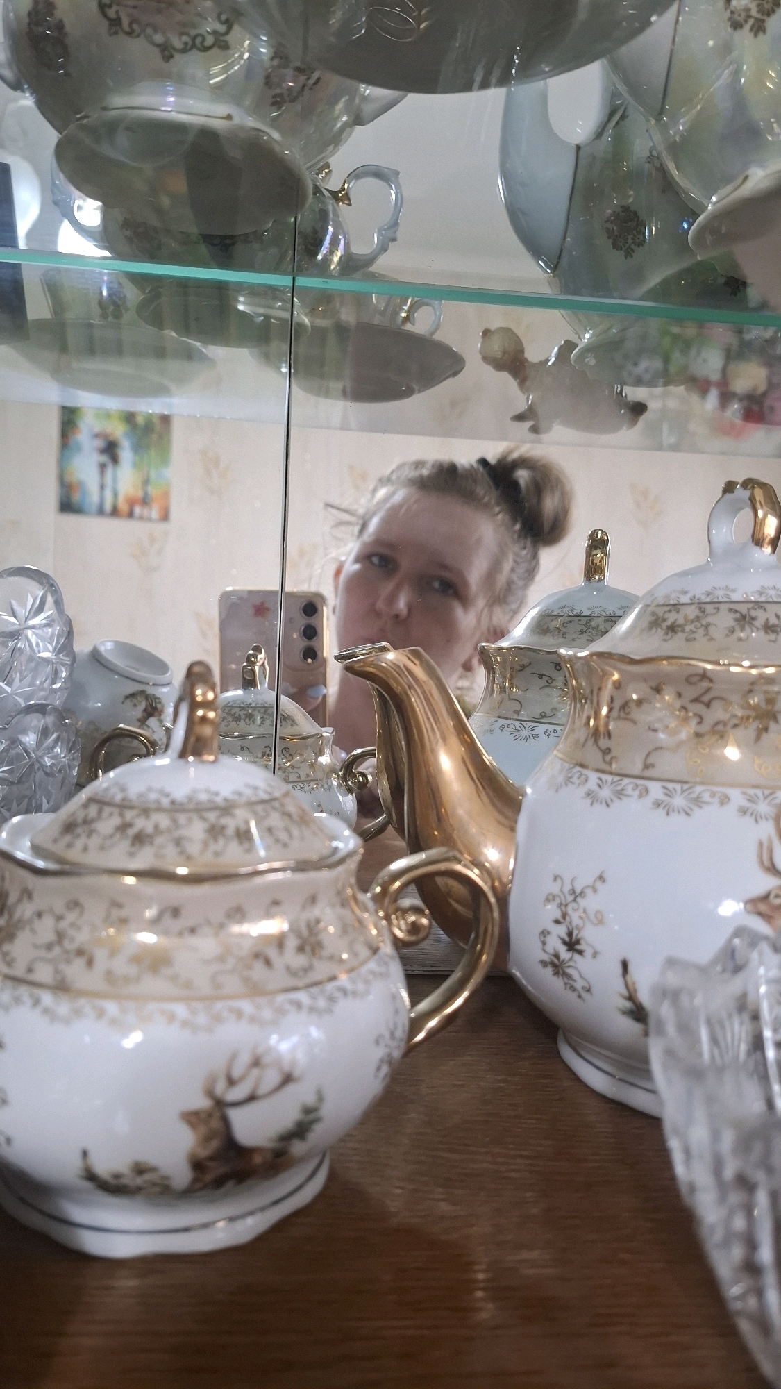 The image depicts a display cabinet filled with ornate porcelain tea sets, predominantly white with gold accents and deer motifs. A woman is visible in a reflection, holding a smartphone and taking a selfie. The cabinet's shelves are made of glass, reflecting the items within and the surrounding environment. The background shows a light-colored, patterned wallpaper and a framed picture on the wall. The scene suggests an indoor setting, likely a home or a shop. There are no discernible landmarks to place this image specifically within Shymkent, Kazakhstan. The focus is on the decorative porcelain ware and the woman's reflection.