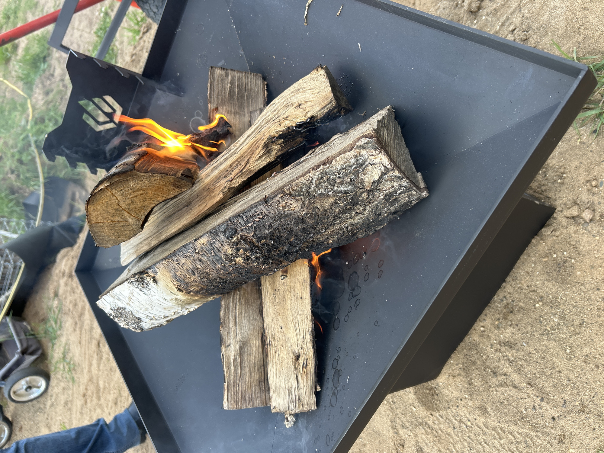 In Chaikovsky, Russia, a close-up shot captures a small fire burning in a black metal fire pit. Several pieces of firewood are arranged within the pit, with flames igniting one of the logs and smoke rising. The fire pit itself is situated on a sandy surface with scattered blades of grass and some pebbles. In the background, out of focus, a portion of a red metal pole and some green foliage are visible, suggesting an outdoor setting. A black metal object with a distinctive logo, possibly part of the fire pit's design or a related accessory, is also visible. The lighting suggests it's daytime, and the weather appears to be mild. The overall scene conveys a sense of a casual outdoor activity, likely a campfire being prepared or maintained.