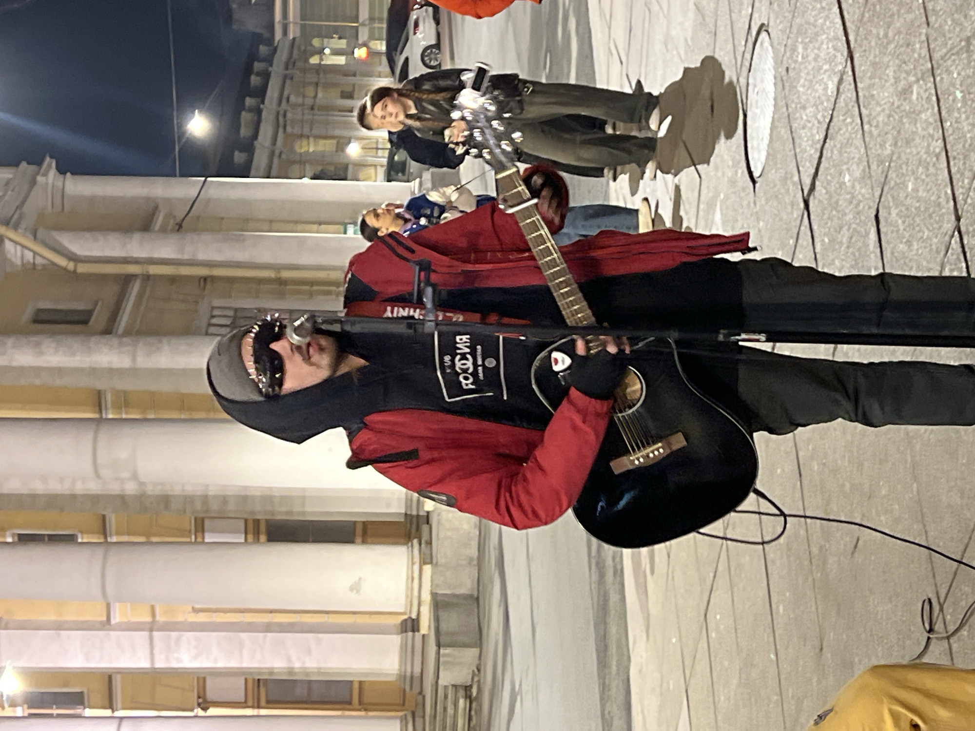 The image depicts a street musician performing in Saint Petersburg, Russia. The musician, wearing a red jacket, black hoodie with white Russian text and logo, sunglasses, and a gray hat, plays a black acoustic guitar into a microphone. He stands on a paved sidewalk next to large, classical-style building with white columns. A small crowd of people are behind him, with one woman holding a smartphone. The ground is gray and patterned. The scene appears to be at night, lit by streetlights.