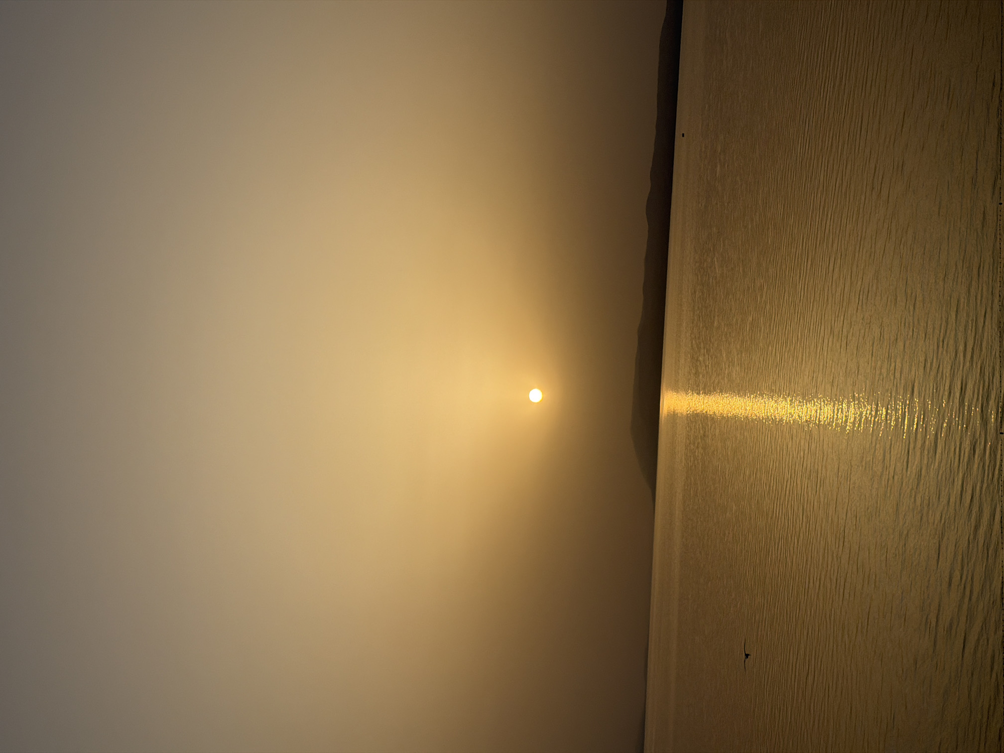 This is a vertical, eye-level shot of a serene sunset over a body of water, likely the Adriatic Sea, as seen from Općina Podgora, Croatia. The sun, a bright, perfectly round orb, is positioned slightly to the left of the image's vertical center and casts a shimmering, golden reflection that stretches across the water. The sky is a gradient of warm, hazy yellows and oranges, transitioning to a muted brown at the top of the frame, suggesting the sun is low on the horizon.
A dark, silhouetted landmass, likely hills or mountains forming the coastline, stretches across the middle distance, a dark contrast to the luminous sky and water. On the water's surface, a small, dark object, possibly a boat or a buoy, is visible near the distant shore. To the left of the frame, a solitary bird is captured in mid-flight, its silhouette stark against the expanse of water and sky. The water itself is textured with gentle ripples, catching the light in undulating patterns. The overall impression is one of tranquil beauty and vastness.