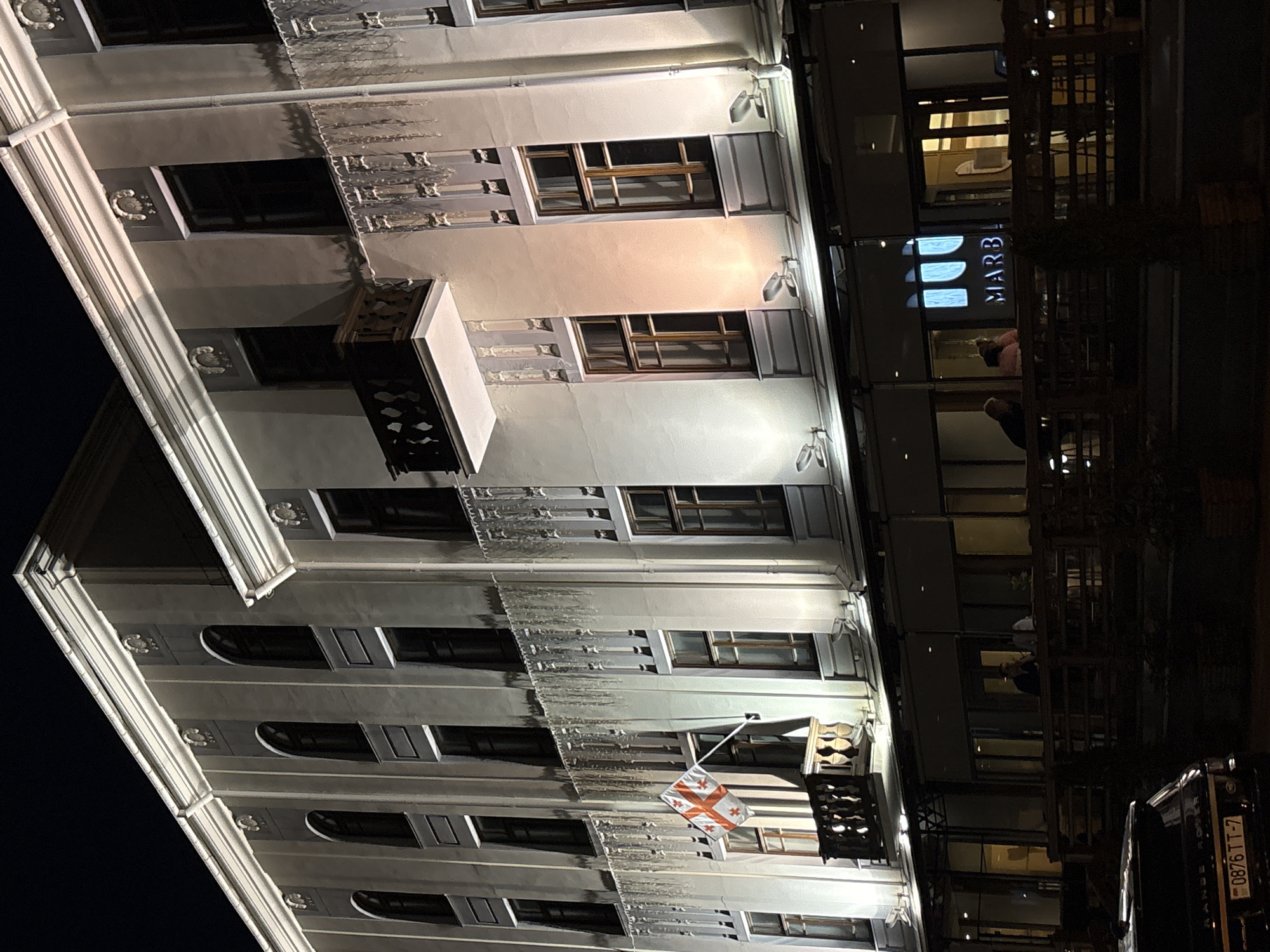 The image depicts a building facade in Minsk, Belarus, at night. The building is multi-storied, with ornate architectural details and a balcony. Georgian flag is displayed on one of the floors. Adjacent to it is a modern glass-walled building with a sign that reads "MARB", and people can be seen inside. A dark-colored Range Rover is parked at the street level, its license plate number "0876 TT-7" is visible.