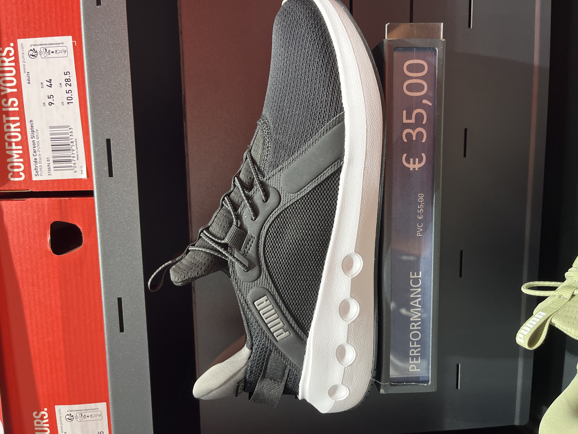 The image depicts a retail display of Puma shoes in what appears to be a store in Rodengo, Italy. In the foreground, a black Puma softstride sneaker with a white sole is prominently displayed on a shelf. To the left of the shoe, a price tag reads "PERFORMANCE € 55,00" and to the right, a larger price tag displays "€ 35,00". Above the shoe, a portion of a red Puma shoe box is visible with text that includes "COMFORT IS YOURS." and a product label for "Softride Carson Sliptech PUMA Black-PUMA White" with size information. The environment is an indoor retail setting, likely a shoe store or a department store with a shoe section. There are no people visible in the image.