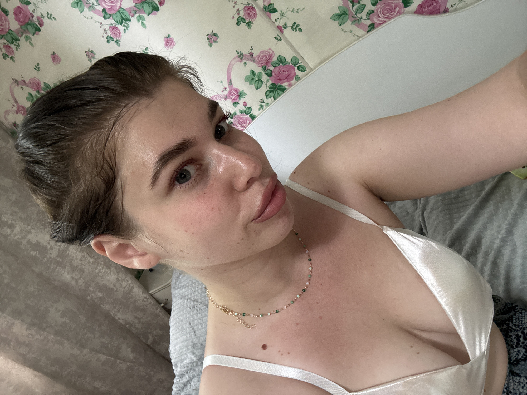 A woman with fair skin, brown hair pulled back from her face, and light blue eyes is taking a selfie indoors. She is wearing a white satin-like camisole with thin straps and a delicate gold necklace. The background includes a white headboard and a wall covered in a floral pattern of pink roses. To the left, there is a light gray fabric with a subtle pattern. The woman is looking at the camera with a slight pout. The location is not discernible from the image.