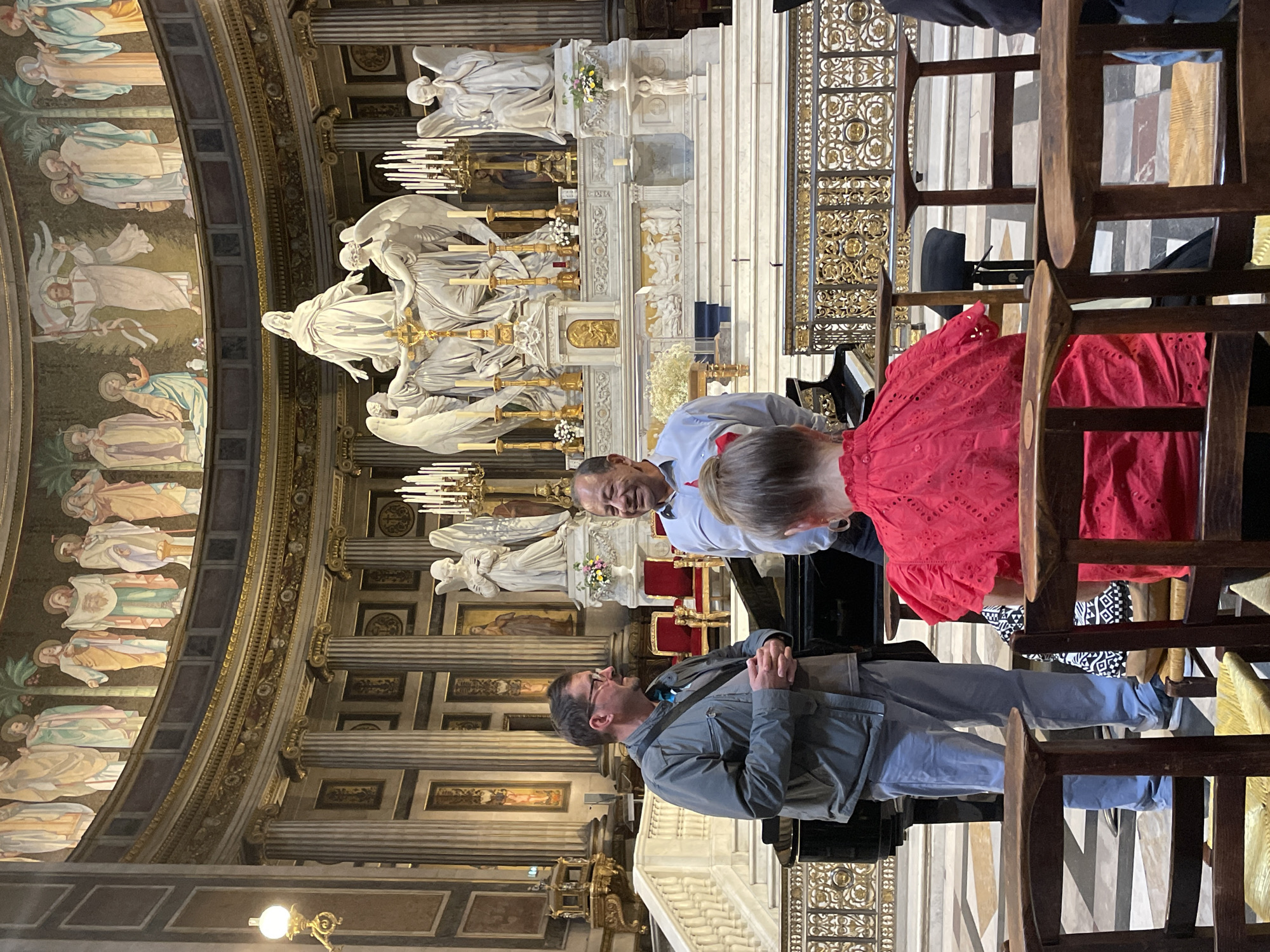 Two men are standing near a grand piano in a large church, possibly a cathedral, in Paris, France. The man on the left wears a grey jacket and light blue pants. He has glasses and is speaking with his hands clasped in front of him. The man on the right wears a light blue collared shirt with a red ribbon tied around his neck. He is smiling and looking towards the man on the left. In the foreground, a woman in a red top is seated, facing away from the camera. The background shows a highly ornate altar area with sculptures and religious artwork, characteristic of Parisian churches like the Panthéon or Notre Dame Cathedral. The architecture includes tall columns and elaborate mosaics.