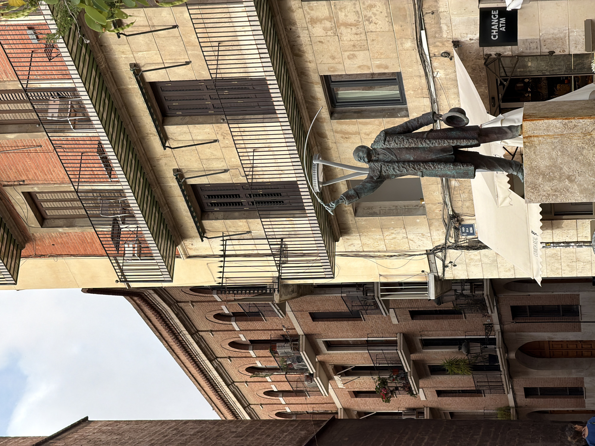 The image shows a statue of a man in a trench coat and hat, holding a whip-like object, standing on a pedestal in front of a building. The building has several balconies with black railings and is constructed with yellow stone on the lower floors and red brick on the upper floors. To the left, another building with red brick and arched windows is visible. A street sign reads "PLAZA DE LA REINA". A sign for "CHANGE ATM" is also visible on the right side of the image. The sky is overcast. The lower left corner of the image shows a person from behind with their back to the camera.