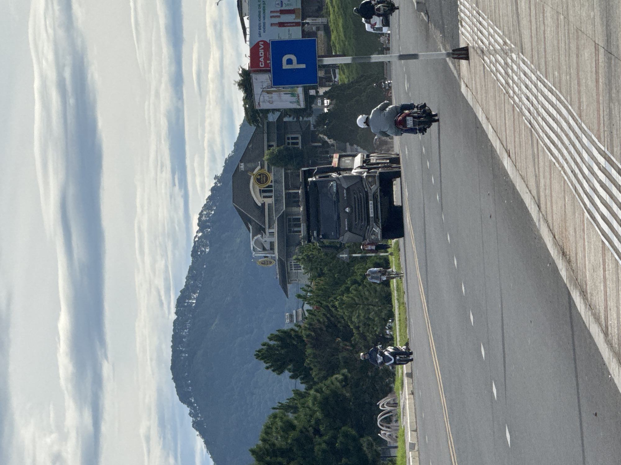 The image depicts a wide, paved road in Dalat, Vietnam, with a mountainous backdrop. On the road, a black dump truck is driving, partially obscuring a large building with a distinctive roofline and several trees in the foreground. To the right of the dump truck, a person on a motorcycle is visible, as well as another person on a bicycle. A blue parking sign with a white "P" is mounted on a pole. The road features yellow lane markings and white dashed lines. A textured sidewalk with tactile paving runs along the right side of the road. The sky is cloudy, with visible cloud formations.