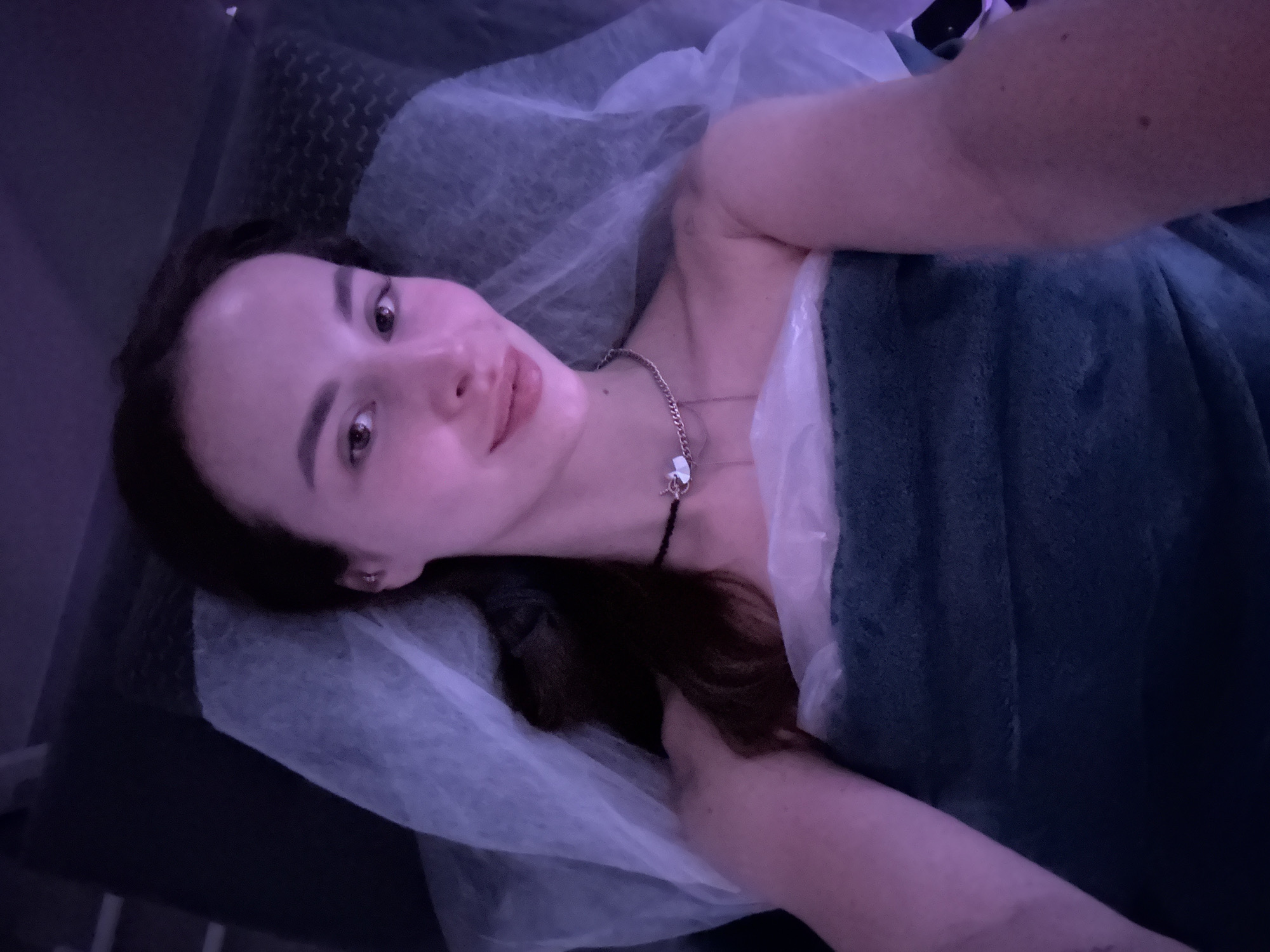 A woman lies on a massage table draped with a white sheet and a blue blanket. She wears a silver necklace with a charm and a black choker. The lighting appears dim, possibly from a spa setting. There is no immediate evidence to confirm the location is Kyiv, Ukraine.