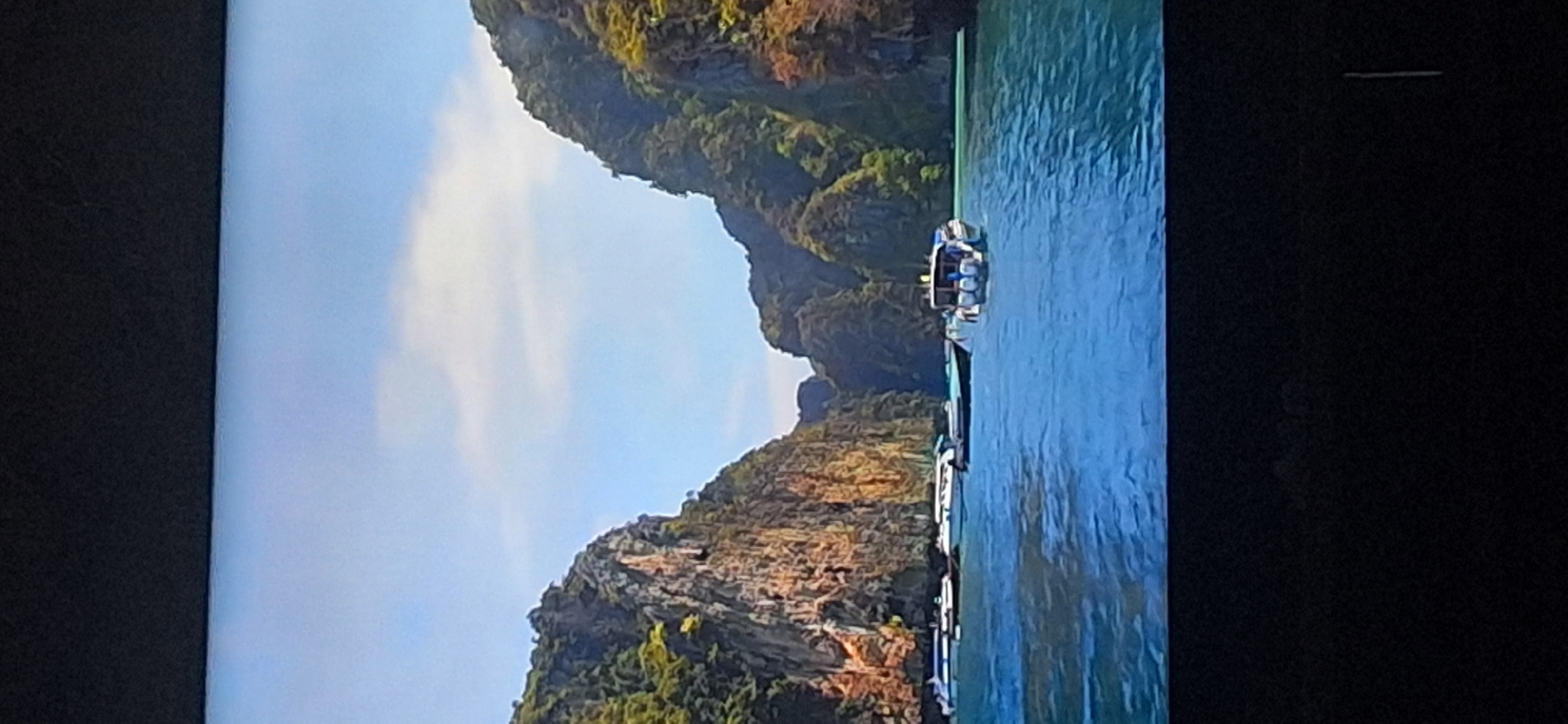 This image depicts a serene natural landscape with towering limestone cliffs covered in lush green vegetation on either side, framing a tranquil blue body of water. Several speedboats are visible on the water, some docked near the shore and others in motion. The sky above is a clear, bright blue with scattered white clouds, suggesting a pleasant day. The scene is one of natural beauty and leisure activity. There is no indication that this image is related to Bila Tserkva, Ukraine; it appears to be a coastal or island location.